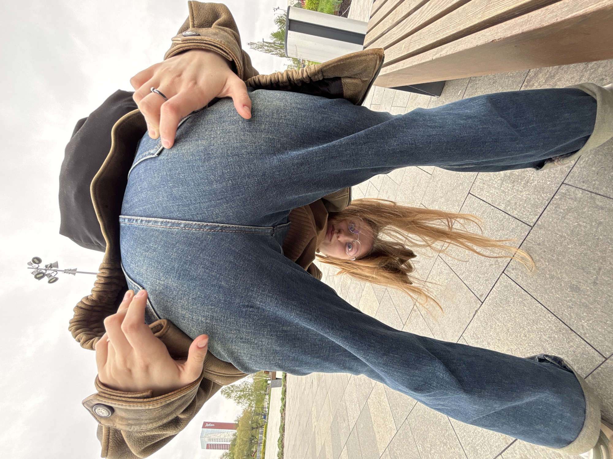 A young woman stands on a paved surface in the city of Perm, Russia, bending over and facing the camera. She is wearing blue jeans, a brown jacket, and glasses. Her long, light-colored hair is falling forward. Her hands are positioned near her rear pockets. A wooden bench is visible behind her. Street lights and a building with the word "Radisson" are in the background. The sky appears overcast.