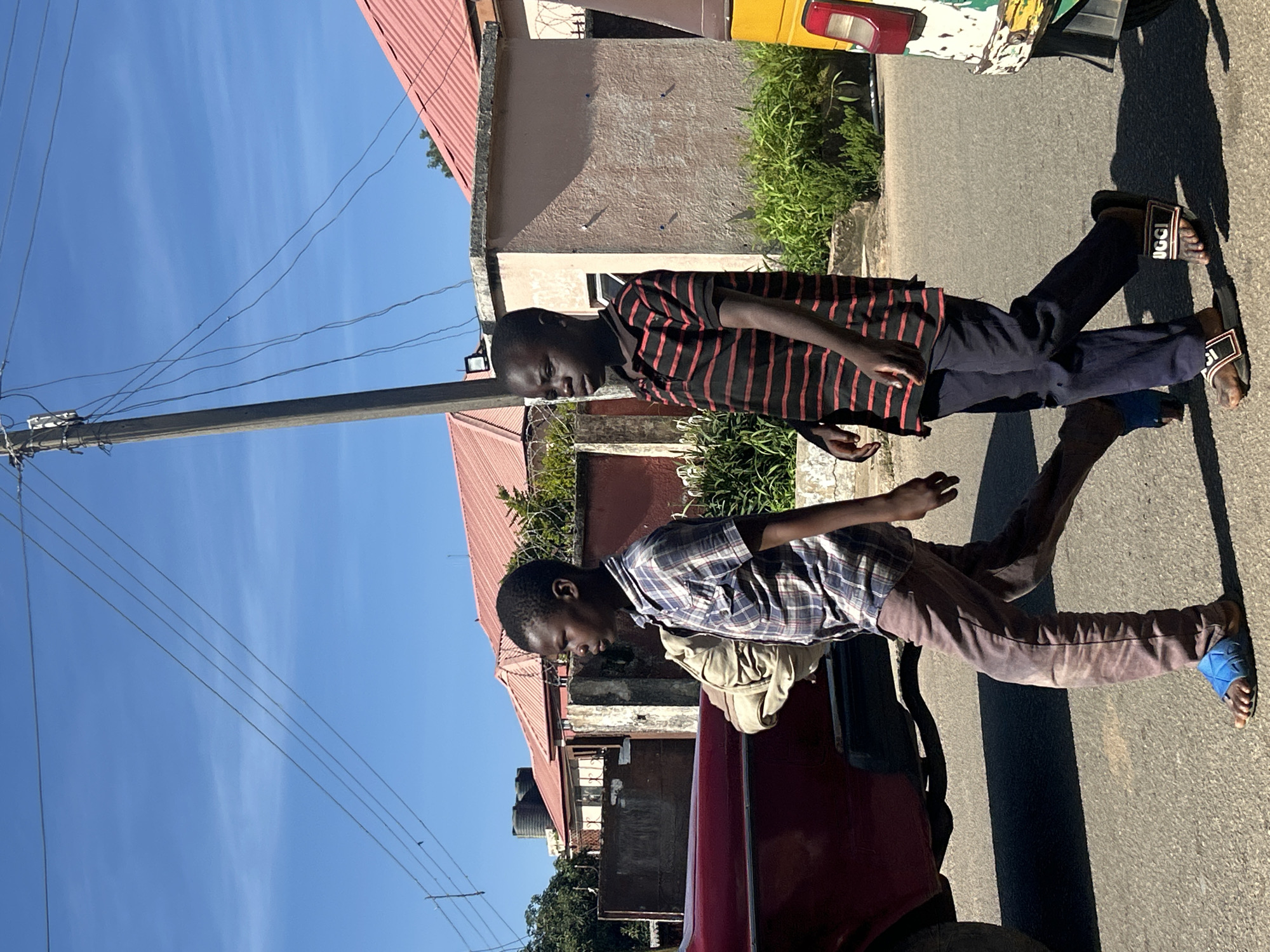 Two young boys are walking on a paved road in Jos, Nigeria. The boy on the left is wearing a blue and white plaid short-sleeved shirt and brown pants. He is carrying a light-colored bag over his left shoulder. He is wearing blue sandals. The boy on the right is wearing a black short-sleeved shirt with red stripes and dark blue pants. He is wearing black slide sandals. They are walking past a red car on the left and a yellow and white vehicle on the right. In the background, there are buildings with red roofs, a concrete wall, and some green foliage. A utility pole with overhead wires is also visible. The sky is clear and blue.