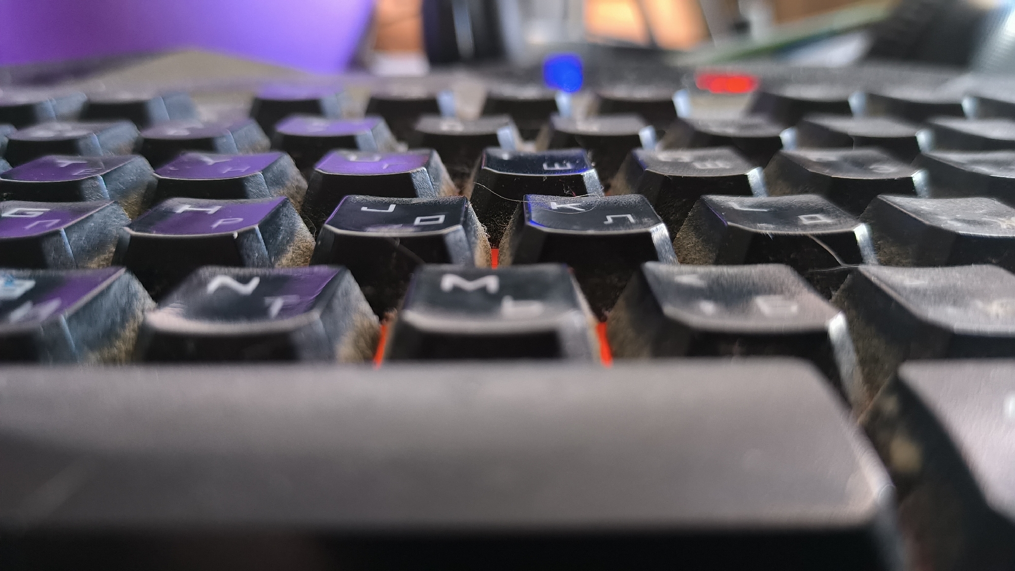 The image is a close-up, low-angle shot of a dusty black keyboard. The focus is on the keys, with some in sharp detail and others blurred due to the shallow depth of field. The keys have a matte black finish, and many have purple symbols or letters on them, though most are too blurry to read. There is visible dust accumulation on the surface of the keys and in the crevices between them. Red and blue lights are visible beneath some of the keys, suggesting it's a backlit keyboard. In the background, a blurry purple object, possibly a monitor or a desk lamp, is visible, along with other out-of-focus elements of what appears to be a desk setup. The lighting suggests it could be indoors, likely in a room with artificial lighting, given the colored backlighting of the keyboard. There is no activity or event taking place; it is a still image of an object. No specific time of day or weather cues are discernible. The location is identified as Kovrov, Russia, but this context is not visually evident in the image itself.