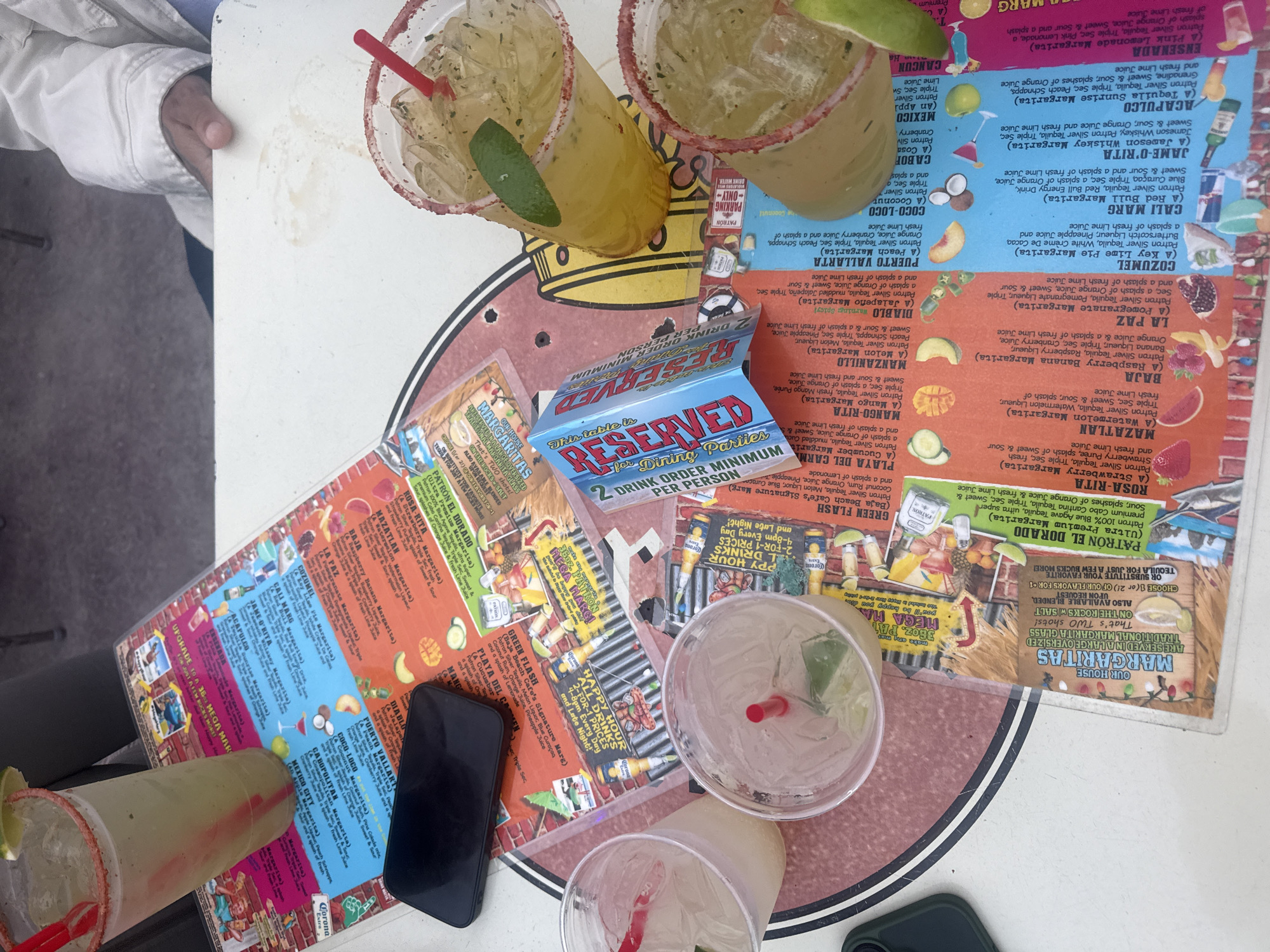 The image captures a table scene in West Hollywood, California, United States. There are several margarita cocktails on the table. Some are full and garnished with lime wedges and rimmed with salt, while others appear nearly empty, containing only ice and straws. A "Reserved" sign is visible, indicating the table is designated for dining parties with a 2-drink order minimum per person. Menus for margaritas are spread across the table, detailing the various types and ingredients. A black smartphone also rests on the table. The table itself features a colorful design with a circular pattern and illustrations of cocktails and other drinks.