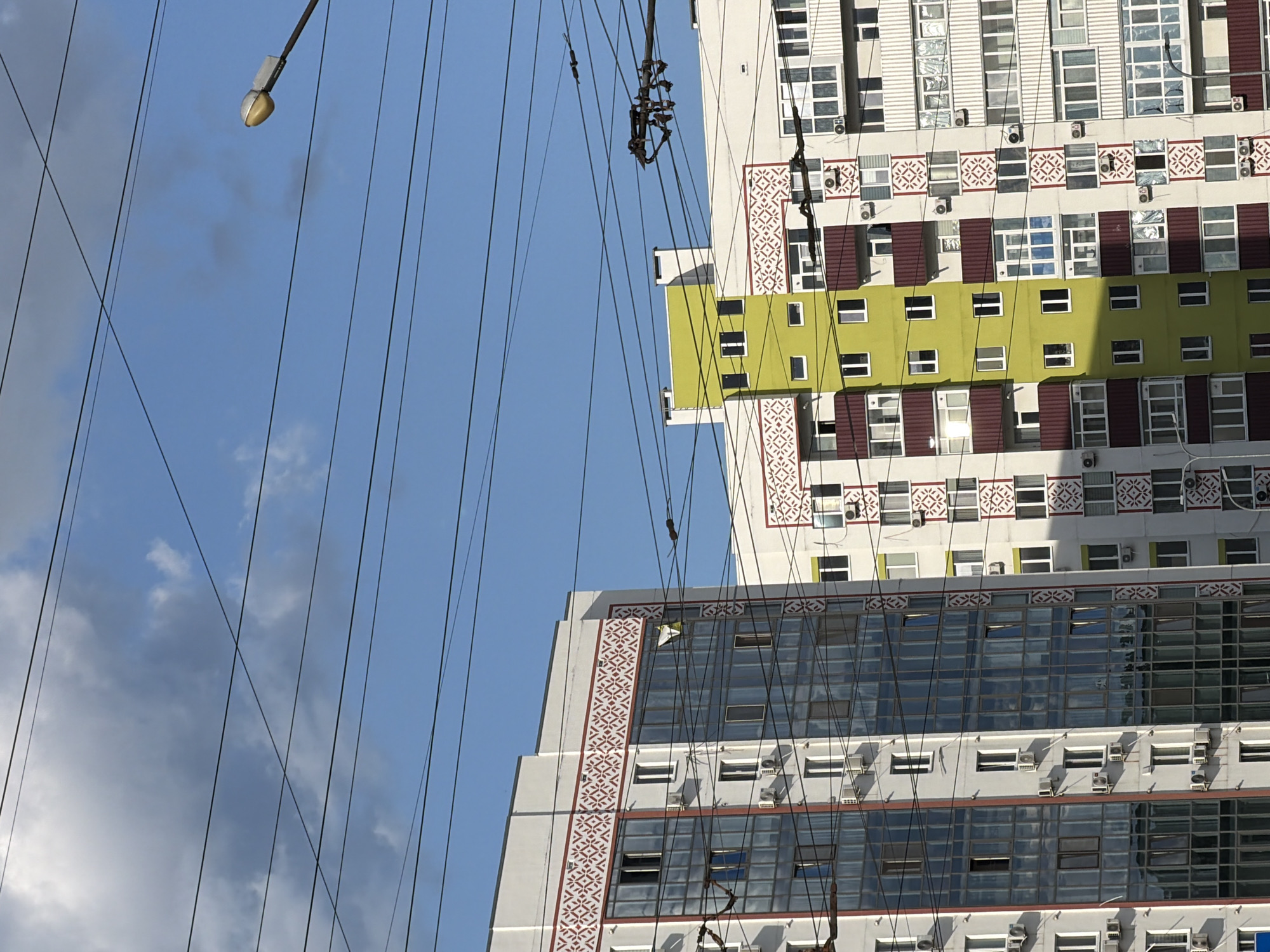 The image shows two modern apartment buildings under a bright blue sky with scattered clouds. A network of overhead wires crisscrosses the sky, and a street lamp is visible on the right side. The building on the left is primarily white with a red and white decorative band near the roofline and red trim around its large, reflective windows. The building on the right has sections painted in bright lime green and white, also featuring the same red and white decorative band. Both buildings are multi-story and appear to be residential, with numerous windows and air conditioning units visible. The scene suggests an urban environment in daylight.