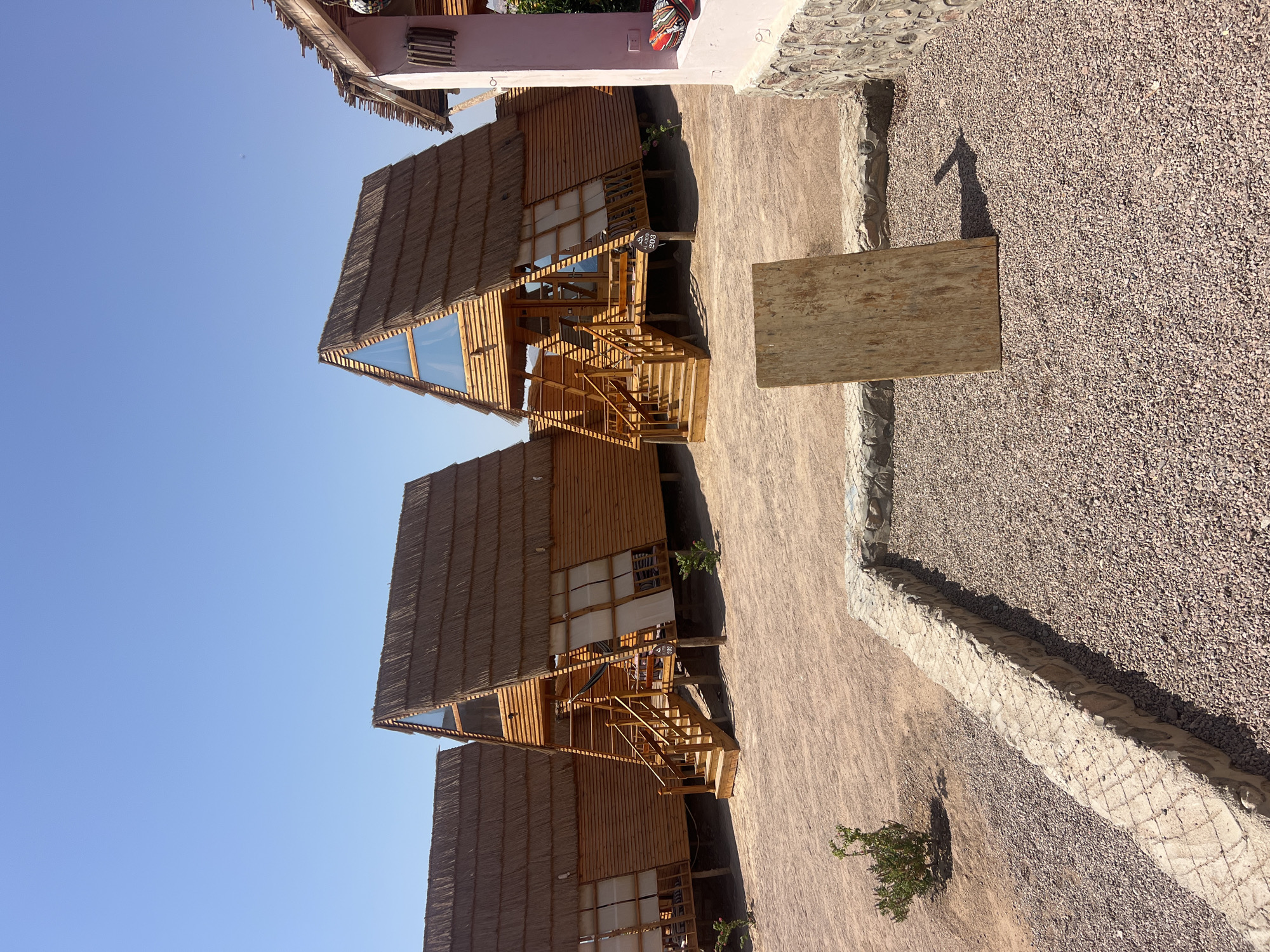 The image shows a row of A-frame cabins with thatched roofs and wooden exteriors, located in Nuweiba, Egypt. The cabins are raised on stilts, with wooden stairs leading up to their entrances. The ground in front of the cabins is covered in sand and gravel, with some small bushes. In the foreground, a wooden sign is propped up in the gravel. The sky is clear and blue. The overall scene suggests a desert resort or campground.