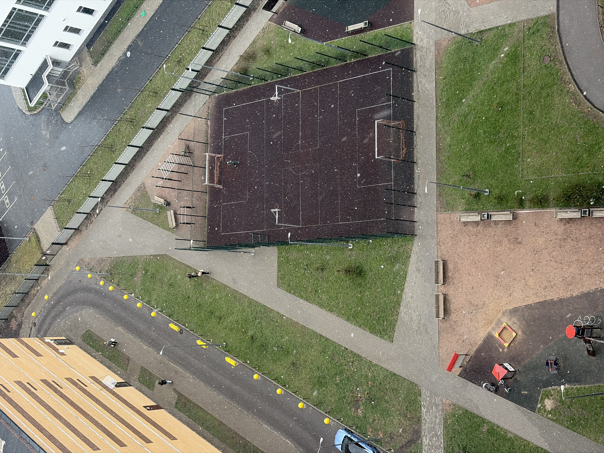 An overhead view captures an urban residential or commercial area in Saint Petersburg during light snowfall. The scene features a mix of paved pathways, green grassy areas, and buildings, under what appears to be daytime conditions, possibly late autumn or early spring given the green grass.
In the center, a dark brown multi-sport court is visible, marked with white lines for football/soccer and basketball. It features two soccer goals (one with a net, one without) and two basketball hoops (one with a net). Several metal posts, possibly for fencing or lighting, surround the court.
To the upper left, a white modern building with large windows and a distinct entrance canopy stands beside a dark paved road. Adjacent to this road is a fenced-off grassy strip.
The lower left features the top of a large, multi-story building with a light brown tiled roof and linear patterns. A curved road with yellow spherical bollards runs along its edge. A blue car is parked on this road.
Various paved pedestrian paths, some light grey and others brownish, crisscross the area, separating green lawns. Numerous wooden benches with metal frames are strategically placed along these paths and near activity zones. Tall, thin streetlights illuminate the pathways.
In the lower right, a children's playground area is visible, featuring a red sandbox and various red and black spring riders or rocking horses on a dark surface.
Several individuals are scattered throughout the scene, walking on pathways or near structures. They appear small due to the high vantage point, generally dressed in dark clothing suitable for cooler weather. No specific activities beyond walking are clearly discernible. The light snowfall adds a speckled texture across all surfaces. No clear text is visible.