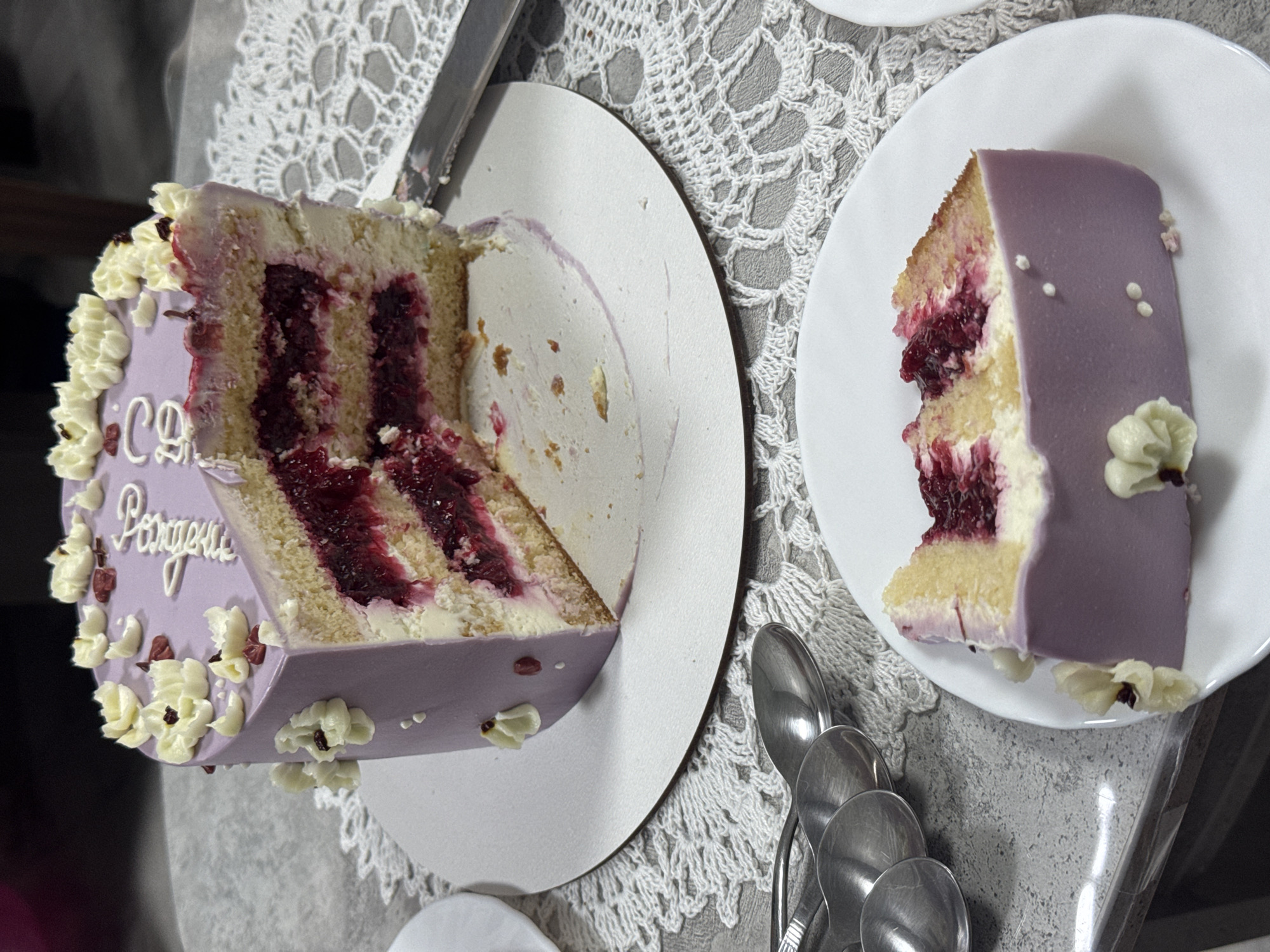 The image depicts a sliced purple cake with a berry filling, resting on a white circular board. The cake is adorned with white floral decorations and edible lettering. A fork is partially visible on the board. A slice of the same cake is presented on a white plate in the foreground, with spoons to the left. The scene appears to be indoors, possibly a home or a bakery, with a table covered by a lace tablecloth. The lighting suggests a natural light source. No specific location in Sosnoviy Bor, Russia, is identifiable from the image.