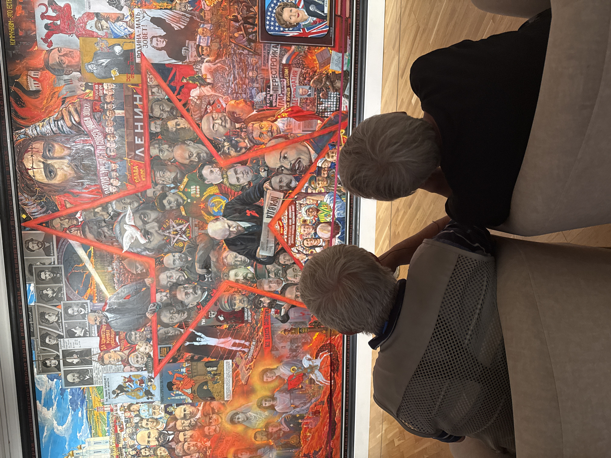 Two individuals are seated in plush chairs, facing away from the camera, observing a large, intricate mural displayed on a wall. The mural is a collage of images, predominantly featuring historical figures, political symbols, and propaganda imagery associated with Soviet history. Prominent elements include portraits of Lenin and Stalin, images of the Red Star, and Russian Orthodox church architecture. Text in Russian is visible throughout the mural. The individuals appear to be elderly, with gray hair. The environment suggests an indoor exhibition space, with a wooden floor visible. The mural itself is framed by a black border. The scene is likely located in the city of Novosibirsk, Russia, given the presence of Soviet-era imagery and the likely context of such a display.
