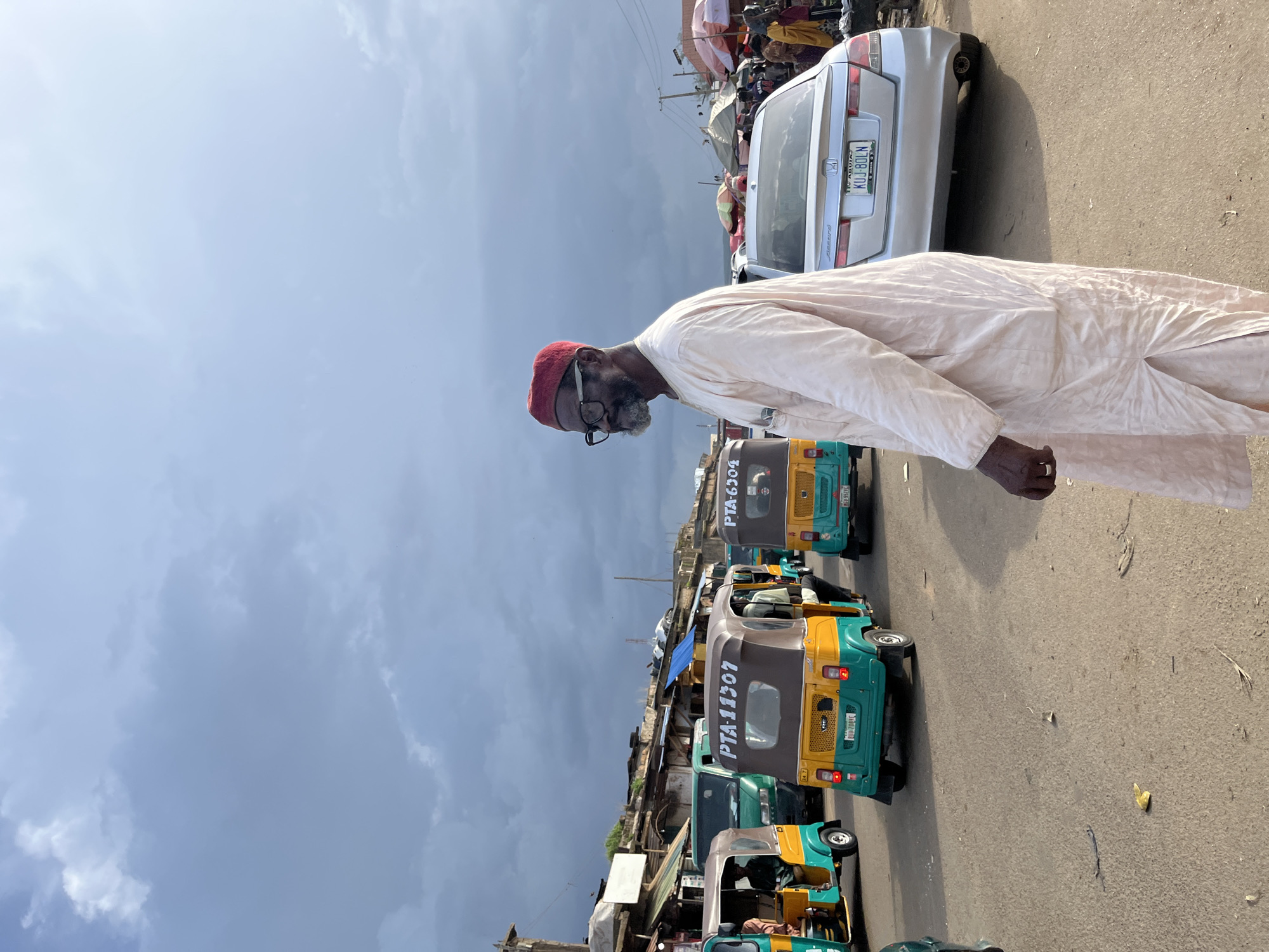 The image depicts a bustling street scene in Rafin Par, Nigeria, under a dramatic, overcast sky. A man in a traditional light-colored robe and a red cap stands in the foreground, facing left and looking away from the camera. He wears glasses and has a beard. Behind him, a silver Honda Accord car with a Nigerian license plate is parked. The street is filled with various vehicles, including several yellow and green auto-rickshaws (also known as Keke NAPEPs) commonly used for public transportation. One auto-rickshaw, with the number PTA-11307 clearly visible, is in the middle ground, and another, PTA-6304, is further back. In the background, market stalls with umbrellas and people browsing can be seen, suggesting a busy commercial area. The overall environment appears to be a typical roadside market in an urban or semi-urban setting in Nigeria.
