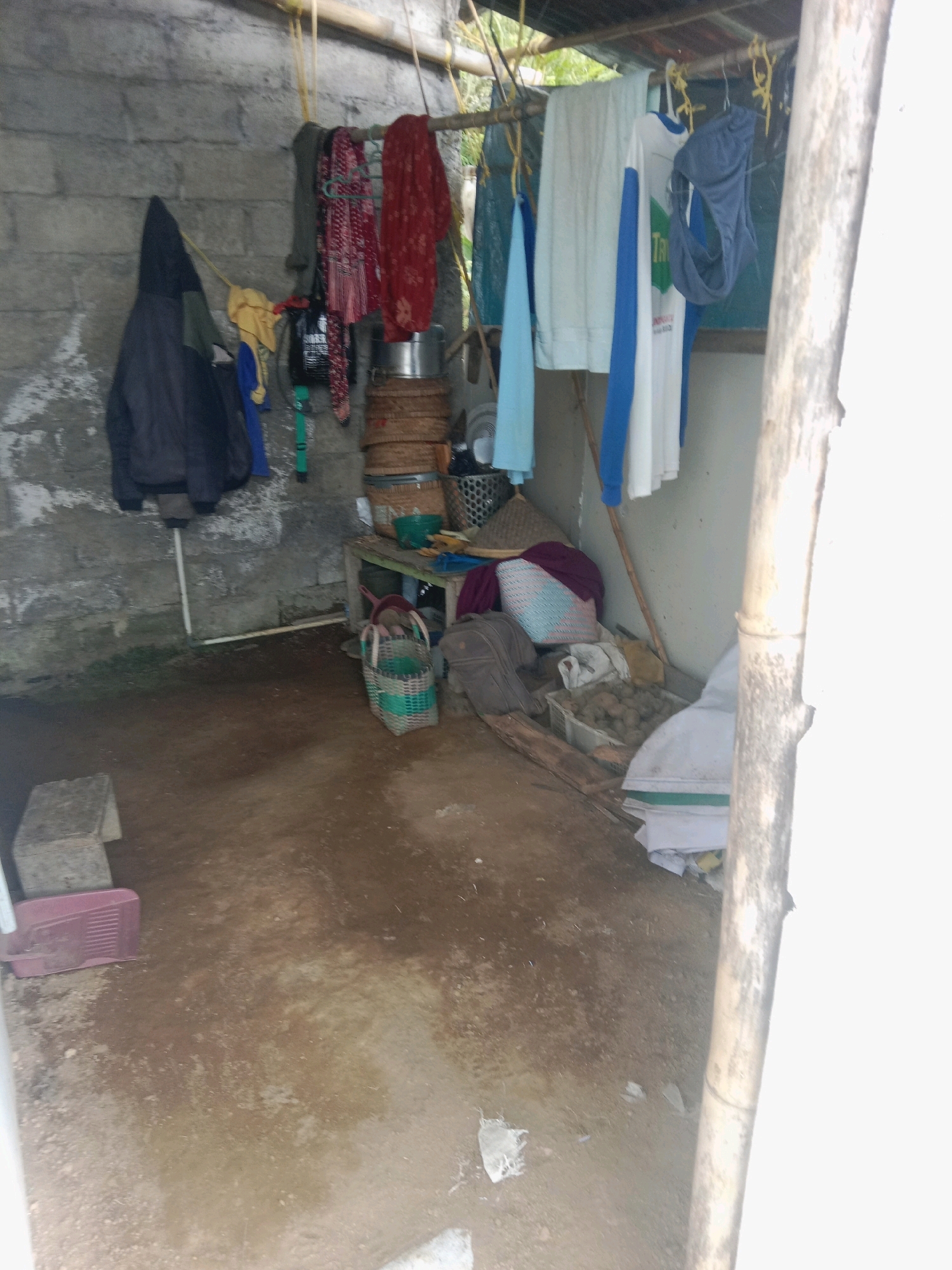 The image depicts a cluttered storage area or lean-to constructed with rough-hewn concrete blocks for walls and a corrugated metal roof supported by bamboo poles. Clothes are hanging on a clothesline that stretches across the top of the space. Among the hanging items are a dark hooded jacket, a yellow garment, a blue garment, a red patterned cloth, and several other pieces of clothing. Below the clothesline, various objects are piled on the floor and on a low wooden table. These include woven baskets, metal pots, a folded blanket, and a white plastic crate filled with potatoes or similar root vegetables. A woven green basket with a handle sits on the floor near the table. To the left of the table, a small, light-colored block is visible, and in the foreground on the left, a pink plastic object, possibly a tray or container, is present. The floor is made of packed dirt, with some wet patches and debris. The scene appears to be outdoors, with glimpses of vegetation visible through the open sides of the structure. The overall impression is one of a functional, albeit disorganized, outdoor storage space in a rural or semi-rural environment.