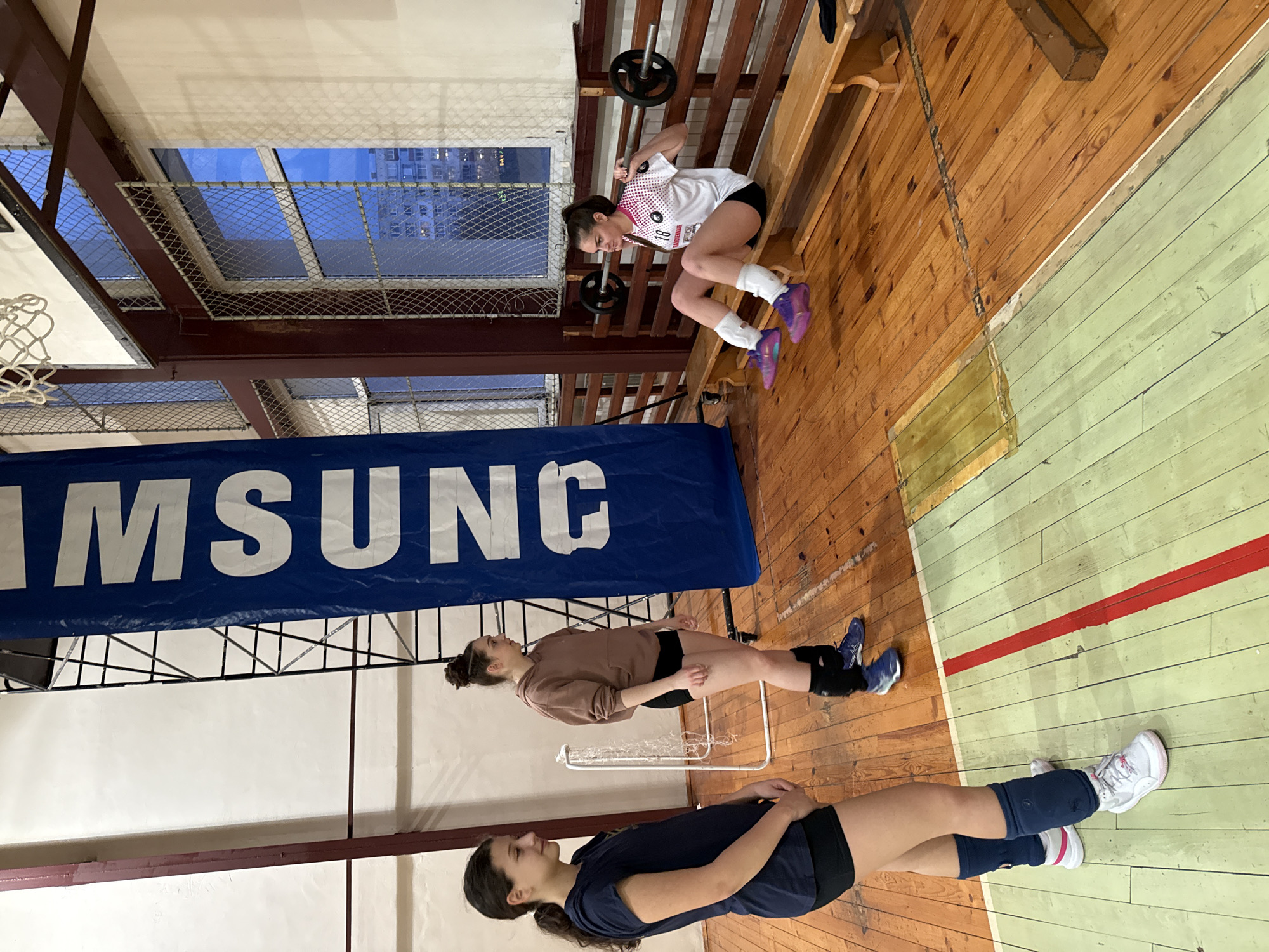 The image shows the interior of a gym, likely in Sofia, Bulgaria, with three young women participating in physical activities.
In the foreground, a woman wearing a dark blue t-shirt and shorts lies on the floor with her knees bent and arms at her sides. The floor is wooden with a painted green and yellow section. Her sneakers are white.
In the middle ground, a woman wearing a brown sweatshirt and shorts sits on a portable chair-like structure. She wears dark knee braces and blue athletic shoes.
In the background, a woman wearing a white and pink volleyball jersey and shorts is performing squats with a barbell on her shoulders. She is positioned in front of a weight bench and wears purple athletic shoes.
A large blue banner with "SAMSUNG" printed in white hangs in the background. Structural elements of the building, including beams and a metal mesh fence over a window, are visible. The window shows a view of buildings in the distance, suggesting an urban setting. The gym's floor is a combination of dark and light-colored wooden planks, with a red line marking a boundary. A basketball hoop is visible in the upper left corner.