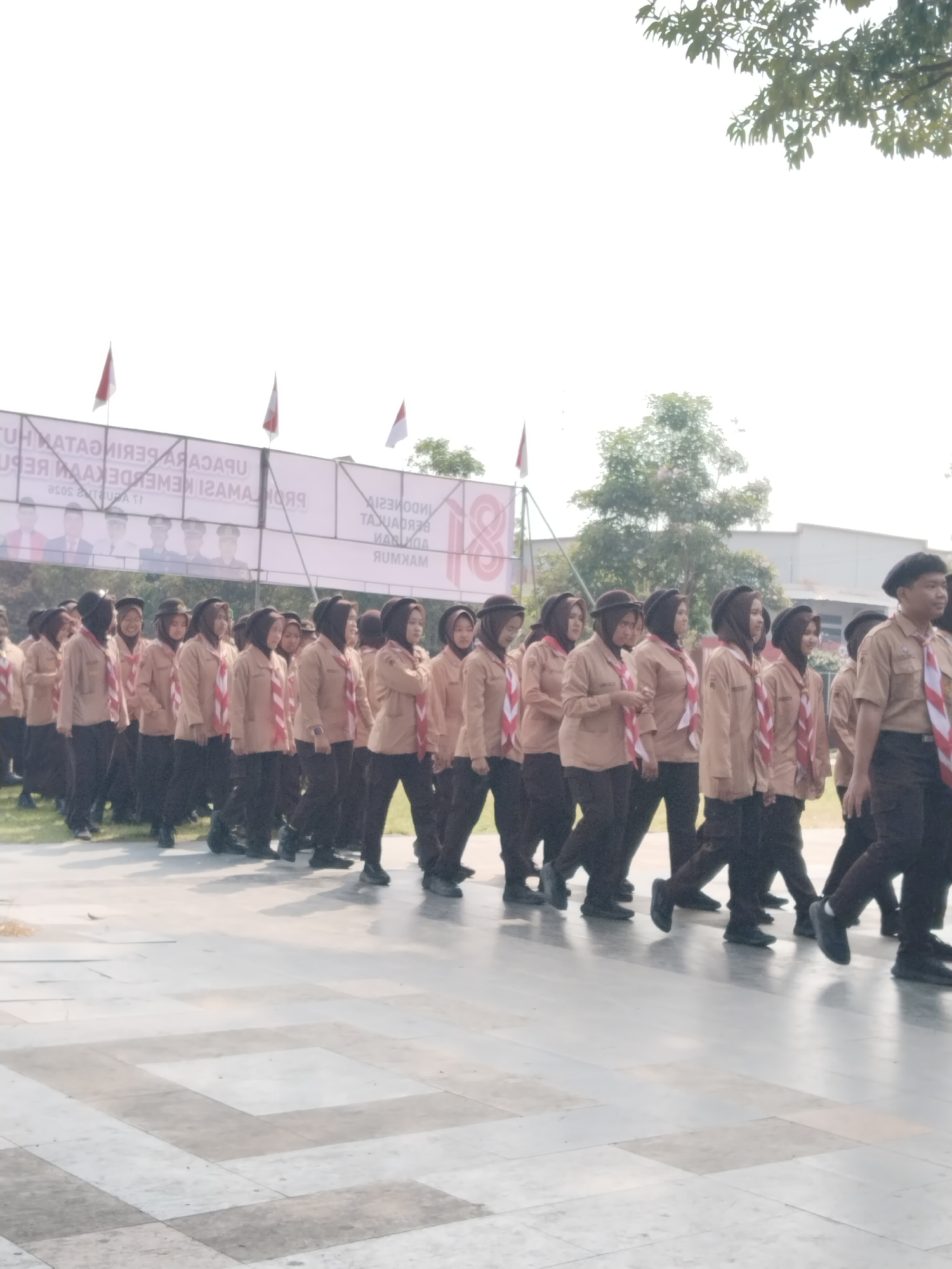 The image shows a group of young people, likely scouts, marching in formation. They are wearing brown uniforms with red and white scarves. The march appears to be taking place outdoors on a paved area, possibly a square or parade ground. In the background, there is a large banner with text that seems to be in Indonesian, and some flags are visible. Trees and buildings are also present in the background, suggesting an urban or semi-urban environment. The overall impression is of an organized event, possibly a ceremonial march. The location is identified as Kecamatan Pati, Indonesia.