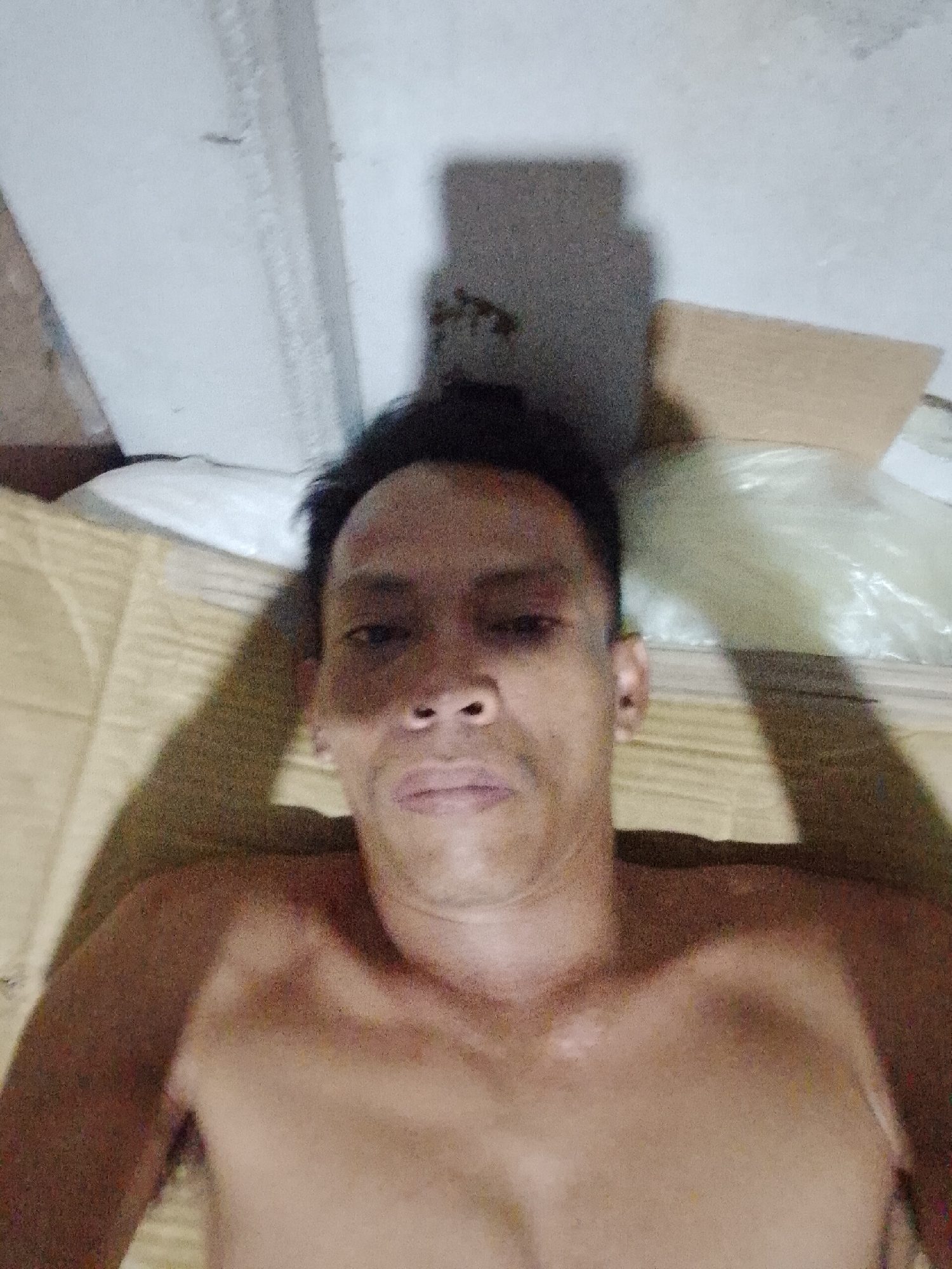 A shirtless man is lying on a tan surface, possibly a mat or blanket. He is facing the camera. Above him, there is a light-colored wall or surface. A cardboard box and a plastic bag are also visible in the background. The location is Bukit Ketok, Indonesia.