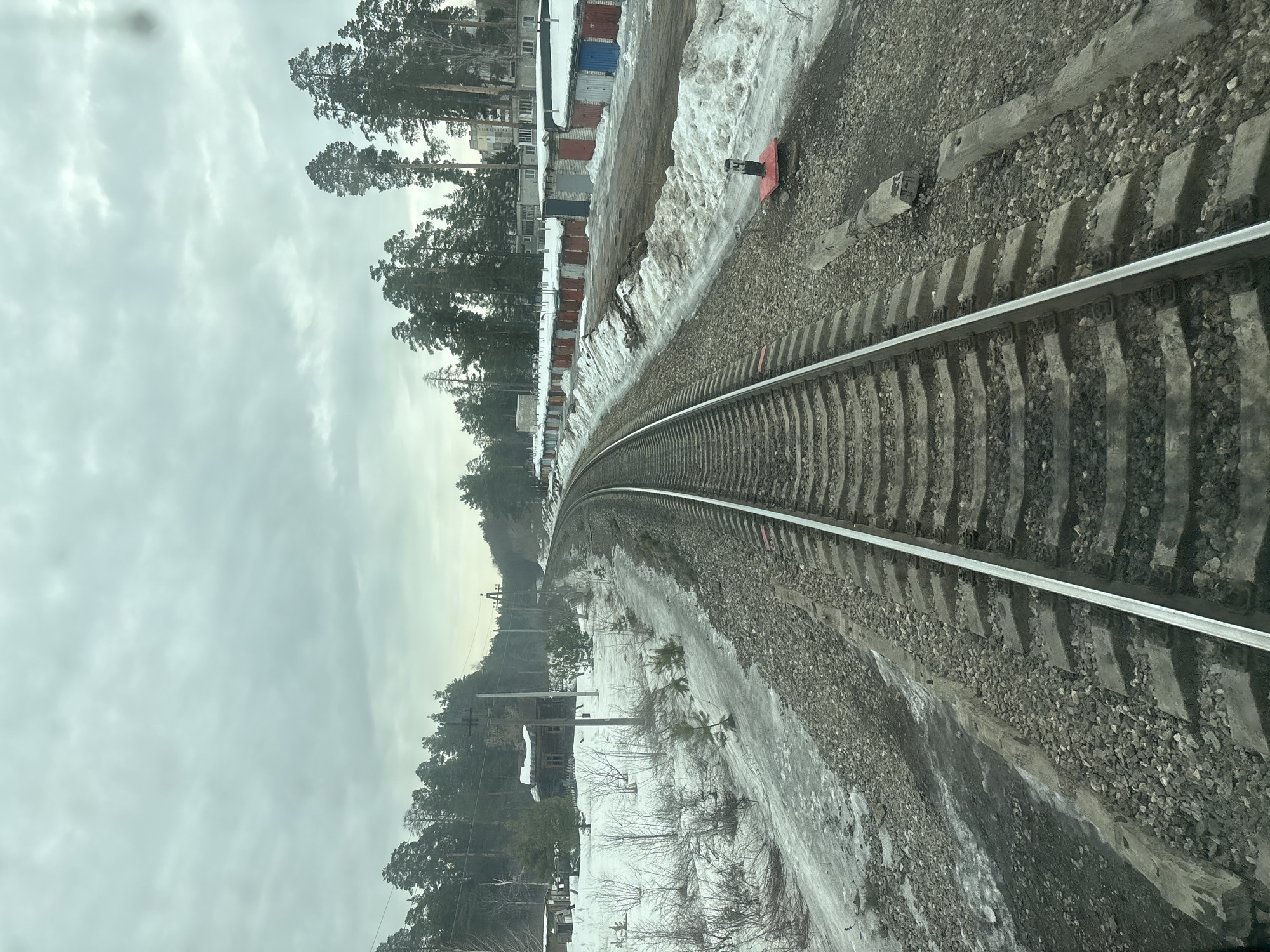 The image, captured in Dimitrovgrad, Russia, depicts a single railway track extending into the distance and curving gently to the right under an overcast grey sky. The track is composed of metal rails secured to concrete sleepers, which rest on a bed of crushed stone ballast. Remnants of melting snow and ice cover the ground adjacent to the track, indicating a late winter or early spring season.
To the left of the railway, the landscape features patchy snow, bare deciduous bushes, and young pine trees. A dense pine forest forms the background on this side, partially obscuring a dark wooden cabin-like structure with snow on its roof, and a smaller, simpler shed to its left. Multiple utility poles carrying overhead lines run parallel to the track.
To the right of the railway, a dirt path is visible, bordered by mounds of snow. Beyond this path, a long series of single-story garages with varied colored doors (including red, blue, and grey) are arranged in a row, their roofs covered with snow. Further behind the garages, several multi-story light-colored buildings are partially visible among tall pine trees. A small, red-based signal or marker post stands near the right rail in the midground. In the foreground, two weathered grey concrete elements lie on the ballast parallel to the right rail. No individuals are present in the scene.
