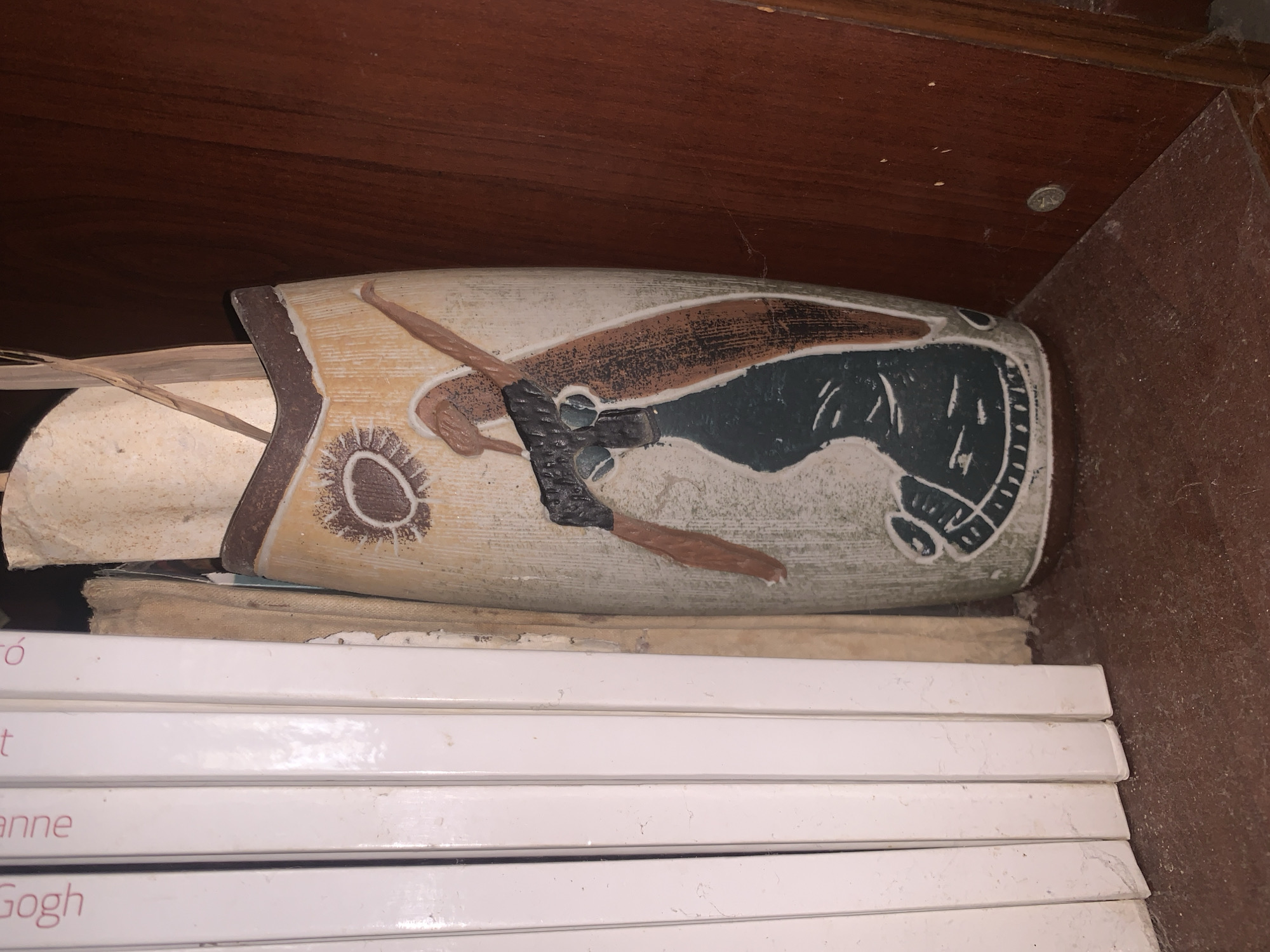 The image shows a decorative vase lying horizontally on top of a stack of books in a dark, enclosed space, possibly a shelf or cabinet. The vase is light-colored with a brown and black design featuring abstract shapes and possibly a stylized figure. The books beneath the vase have white spines with text visible on the bottom spine reading "Gogh." The environment appears dusty with visible cobwebs in the corners of the shelf. The image location is not provided and cannot be determined from the visual content alone.