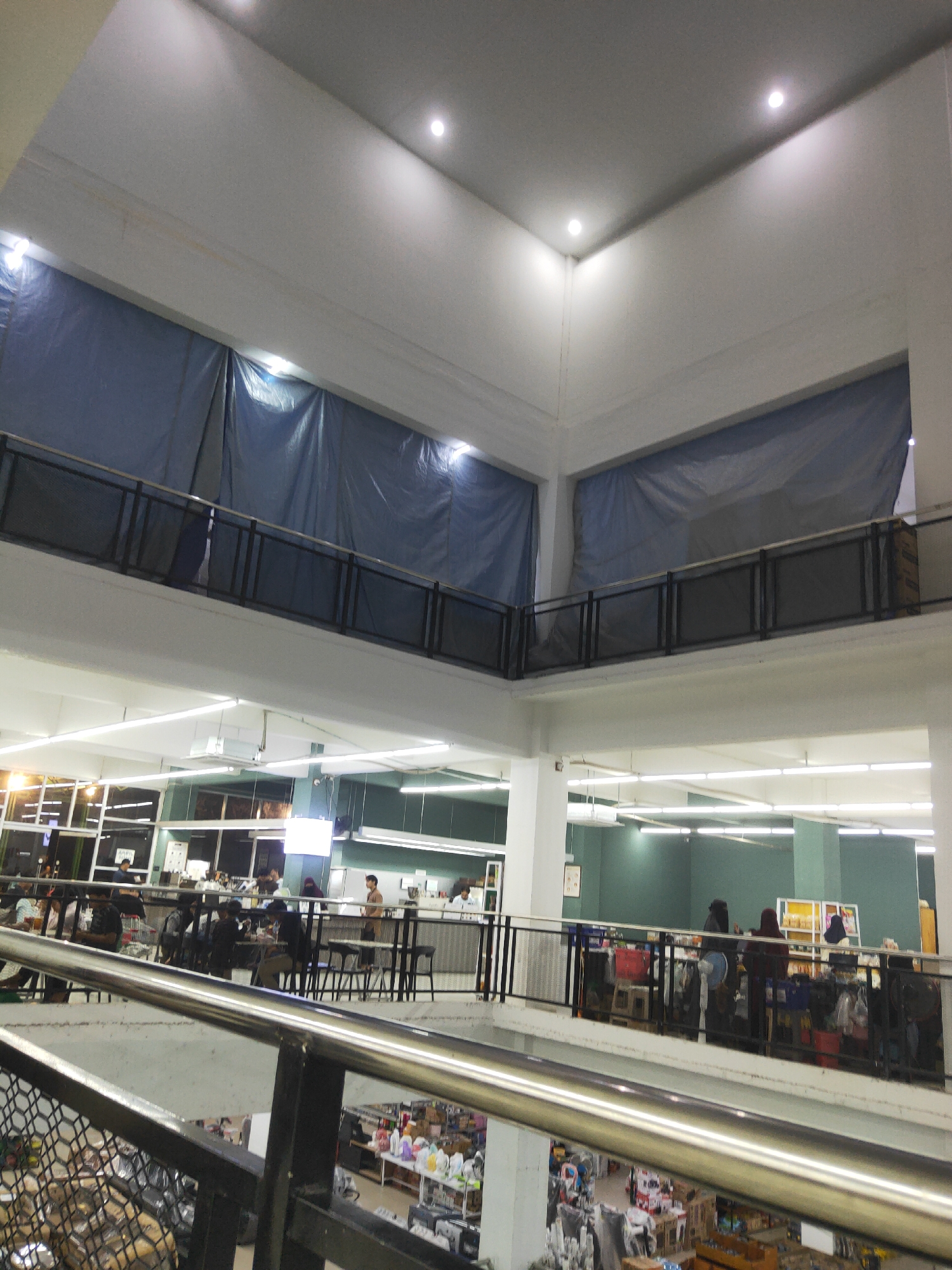 The image shows the interior of a multi-level building, possibly a mall or department store, in Sintang, Indonesia. The foreground features a metal railing with a mesh section, suggesting a balcony or upper-level walkway. In the mid-ground, a second-level walkway with a similar railing overlooks a lower level. People are visible on the second level, some seated at tables, possibly in a food court area or cafe. The walls of the upper levels are covered with large blue tarpaulins. The lower level is filled with shelves of merchandise, indicating a retail space. The lighting appears to be a mix of overhead fluorescent lights and spotlights.