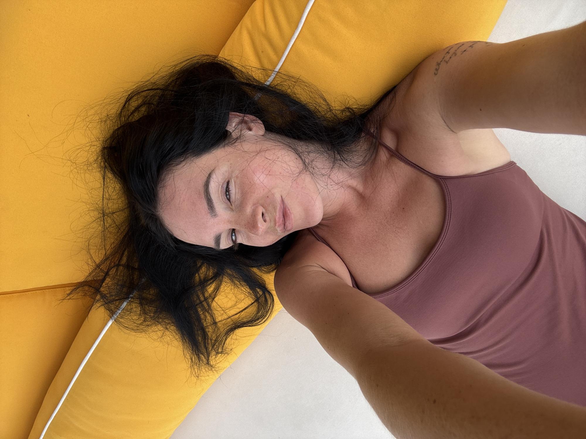 A woman is lying on a yellow surface, likely a sun lounger or cushion, with white piping along the edge. She has long, dark, messy hair spread out around her head. Her head is tilted slightly to her right, and she is looking at the camera with a playful expression, one eye squinting and her lips pursed. She is wearing a mauve-colored camisole. Her right arm is extended outwards, towards the camera, and on her upper right arm, a tattoo is visible with some writing. The background appears to be a light-colored surface, possibly a deck or towel. The scene captures a relaxed and intimate moment, likely taken as a selfie. The location is identified as North Kuta, Indonesia, suggesting an outdoor, possibly coastal or poolside setting given the type of furniture and the woman's attire.