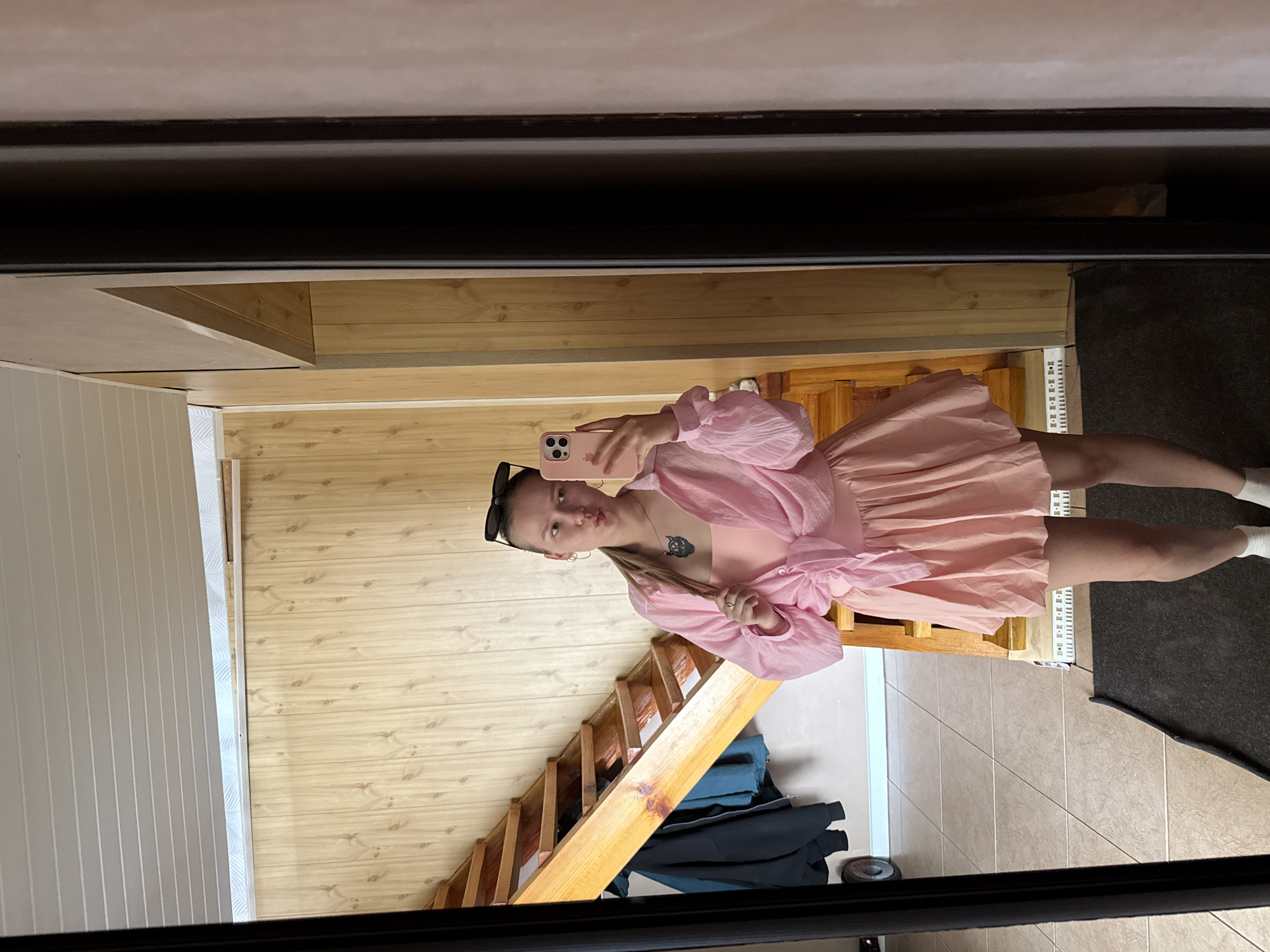 A young woman is taking a selfie in a mirror, wearing a light pink dress with puffy sleeves. She is holding a pink phone. There is a wooden staircase in the background, and a mix of wood paneling and white paneling on the walls. Some clothing is piled beneath the staircase. The floor is tiled and part is covered with a dark rug. The image shows a mirror frame, suggesting the setting is indoors, possibly a home interior. I cannot determine whether or not the location is in Zhigalki, Russia.