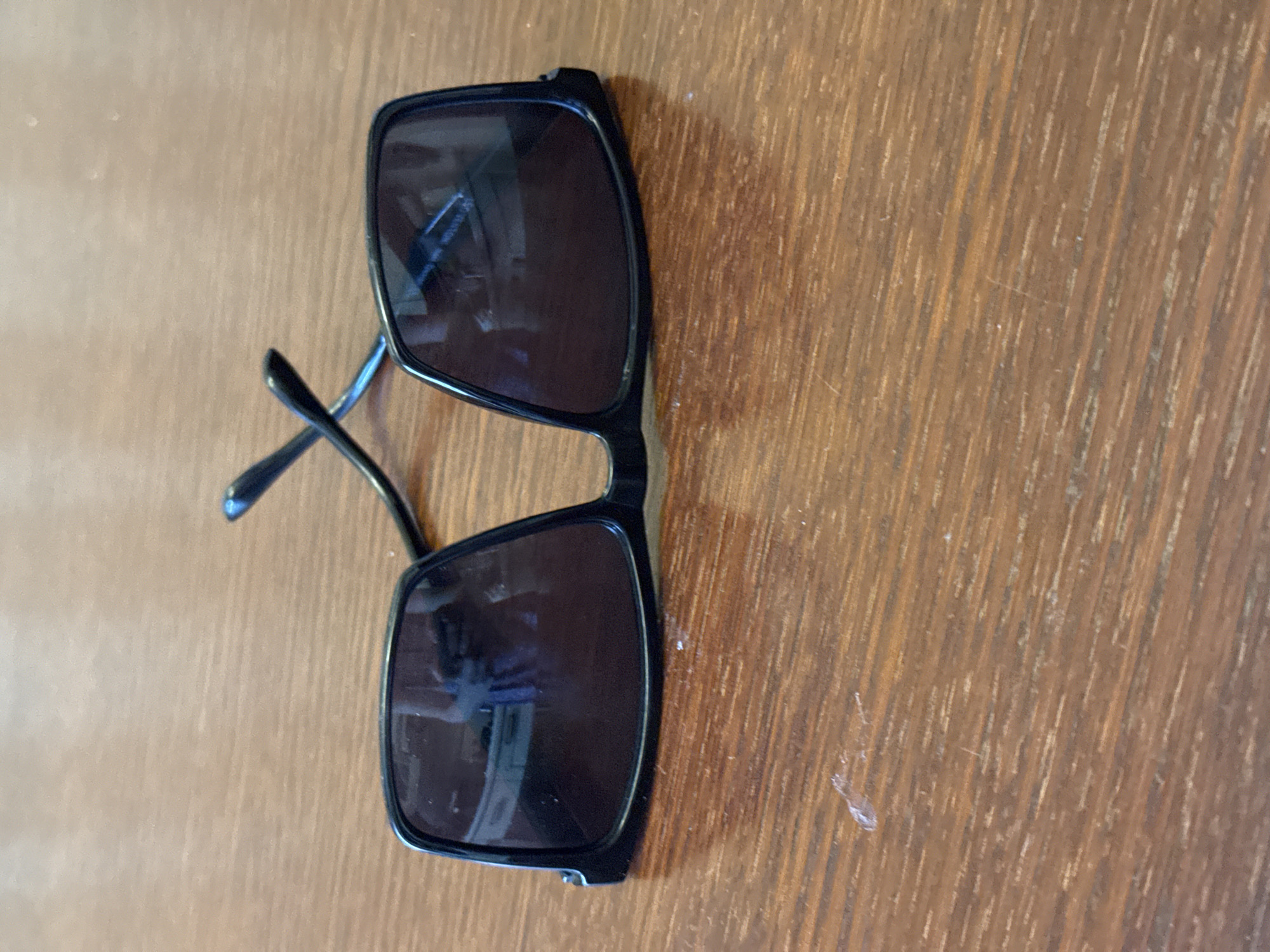 A pair of black-framed sunglasses rests on a wooden surface. The sunglasses are oriented vertically, with the lenses facing upwards. The wooden surface has a visible grain pattern and appears to be a tabletop or desk. The lighting suggests an indoor setting, with a shadow cast by the sunglasses, but no specific time of day can be determined. There are no people or activities depicted in the image. The lenses of the sunglasses are dark and reflective, obscuring any view through them. One of the arms of the sunglasses is slightly extended. There is no visible text on the sunglasses or the surface.