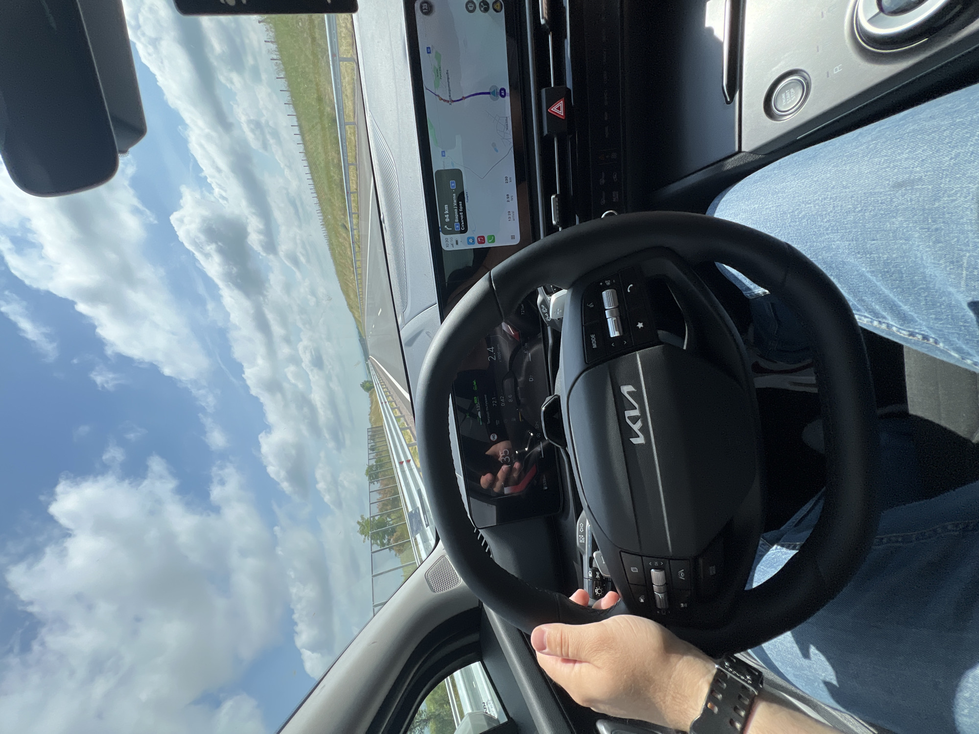 The image captures the interior of a modern car, likely a Kia due to the prominent logo on the steering wheel, as it travels on a highway under a bright, partly cloudy sky. The perspective is from the driver's seat, showing the driver's right hand holding the steering wheel and their light blue jeans.
**Subjects and Objects:** The main subjects are the car's interior components and the road ahead. Key objects include the black leather-wrapped steering wheel with the "Kia" logo and control buttons (one labeled "MODE"), a widescreen digital dashboard and infotainment system, and a "START STOP" engine button on the right side of the dashboard. Outside the car, a multi-lane road, guard rails, grassy verges, and a distant tree line are visible under a sky dotted with white clouds. A wristwatch is visible on the driver's wrist.
**Activity:** The scene depicts the activity of driving a car, evidenced by the driver's engaged posture, the active navigation system, and the moving road environment.
**Notable Details:** The time of day is daylight, with clear visibility. The weather is partly cloudy. The infotainment screen displays navigation information, showing a route with "94 km" remaining to a destination, and listing "Варна / Varna" and "Слънчев бряг" (Sunny Beach) as key locations. The screen also indicates a current time of "13:29", an estimated remaining travel time of "2:59", a total remaining distance of "230" km, and an estimated arrival time of "16:33". The digital instrument cluster, partially visible, shows readings such as "72.1", "0:42", "86", "2.4 x 1000 rpm" (2400 RPM), and "D7" (indicating the car is in 7th gear, likely automatic). Several speed-related numbers are also visible in the cluster or its reflection, including "133", "140", and "1356". A red hazard light button is visible on the center console. The visible locations confirm the setting is in Bulgaria, in the region of Chokoba. No specific emotions are discernible.