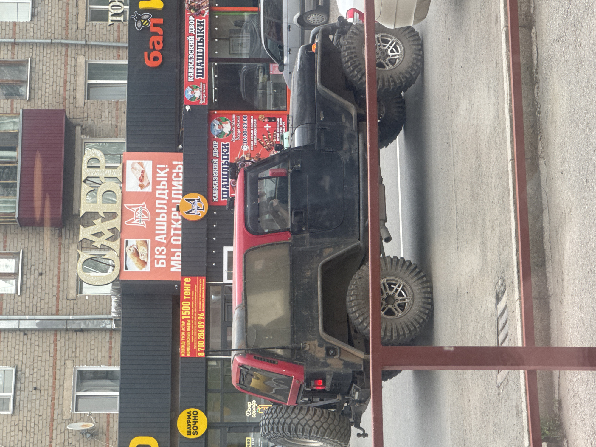 This media file shows a side view of a black and red off-road vehicle parked on a paved surface, possibly a street or parking lot. The vehicle has large, rugged tires. Behind the vehicle, there is a multi-story building with a brick facade and several windows.
Several signs are visible on the building. One large sign, partially obscured, appears to be for a business called "MEGALODON" with images of food. Another prominent sign in red and white text, repeated twice, reads "КАВКАЗСКИЙ ДВОР ШАШЛЫКИ" (Caucasian Courtyard Shashlik). This sign also includes details about the business being "always fresh" and its operating hours, "10.00-23.00". There are also signs with Russian text, including one that says "БІЗ АШЫЛДЫҚ! МЫ ОТКРЫЛИСЬ!" (We are open!). Another sign displays a phone number "8 700 286 09 96" and mentions "1500 тенге" for "КЕШЕНДІ ТҮСКІ АСТАР" (Complex Lunch). A yellow circular sign reads "ШАУРМА СОЧНО" (Shawarma Juicy).
The lighting suggests it is daytime. The scene appears to be in a commercial area, with the vehicle likely parked in front of the businesses. The overall impression is of an urban or semi-urban setting.