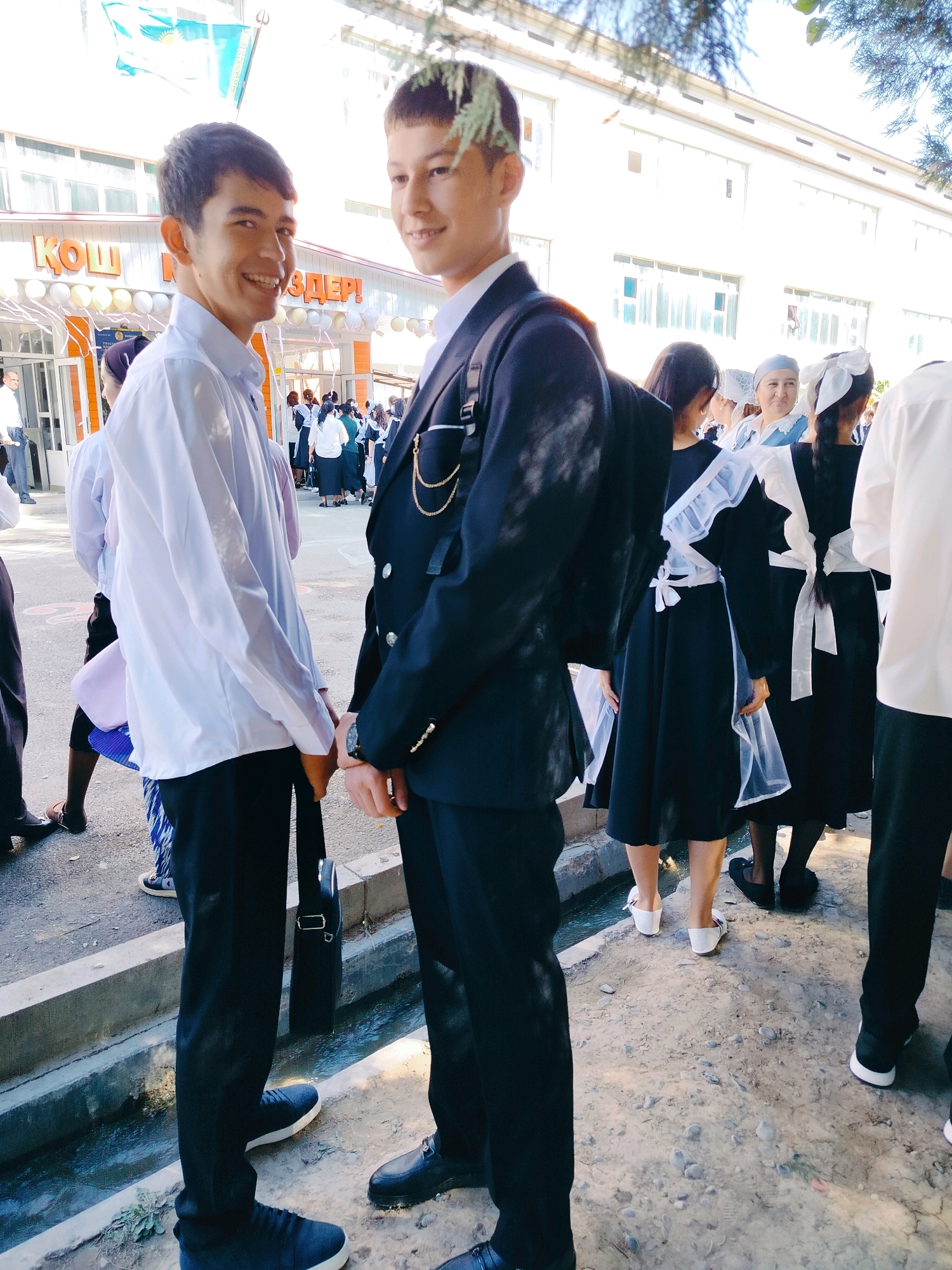Two young men, dressed in formal attire, stand facing forward in a courtyard. The man on the left wears a light blue collared shirt and black pants, while the man on the right is in a dark suit with a white shirt. Both have dark hair. Behind them, a group of young women in traditional school uniforms, consisting of black dresses with white aprons and collars, are visible. The background shows a building with windows, decorated with balloons and streamers, suggesting a school event. The ground is paved and there is a concrete ditch running along the bottom of the image. A flag of Kazakhstan is visible in the upper left corner. The scene appears to be in the city of Kölkent, Kazakhstan, possibly during a school celebration or commencement ceremony.
