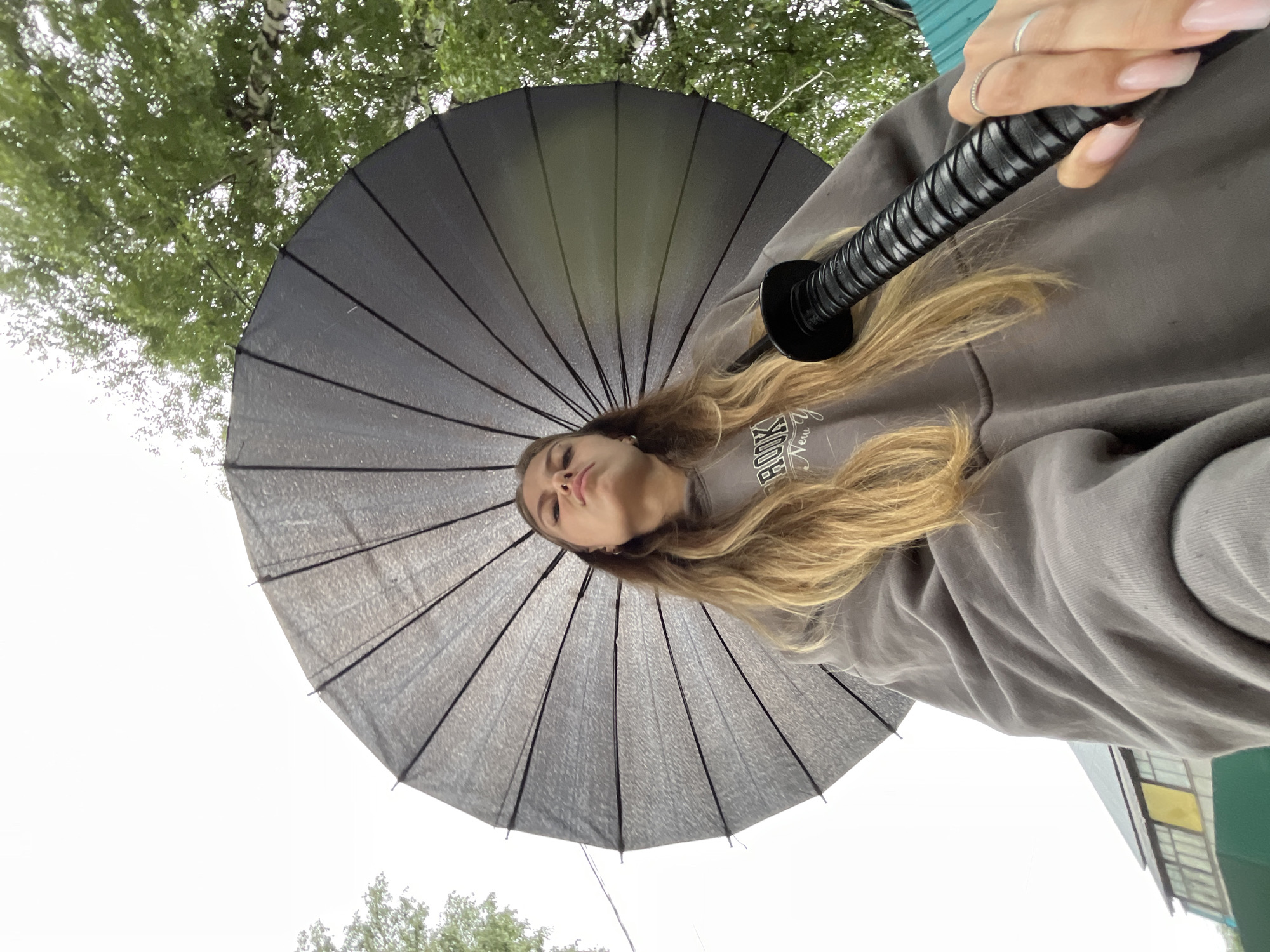 In an outdoor, low-angle shot, a young woman is positioned beneath a large, dark umbrella. The umbrella's spokes are visible, fanning out around her. She has long, blonde hair and is looking upwards with a serious expression. She is wearing a gray hooded sweatshirt with the words "BROOKLYN NEW Y" visible on it. Her right hand, adorned with a ring, is holding the black, ribbed handle of an object, which appears to be the handle of a katana or similar sword. In the background, lush green trees are visible, as well as a building with a multi-colored facade. The sky is overcast and bright, suggesting a cloudy day. The overall scene conveys a sense of dramatic pose or preparation, possibly for a photo shoot or artistic representation. No specific location within the city of Фоки, Russia can be determined from the visual information presented in the image.