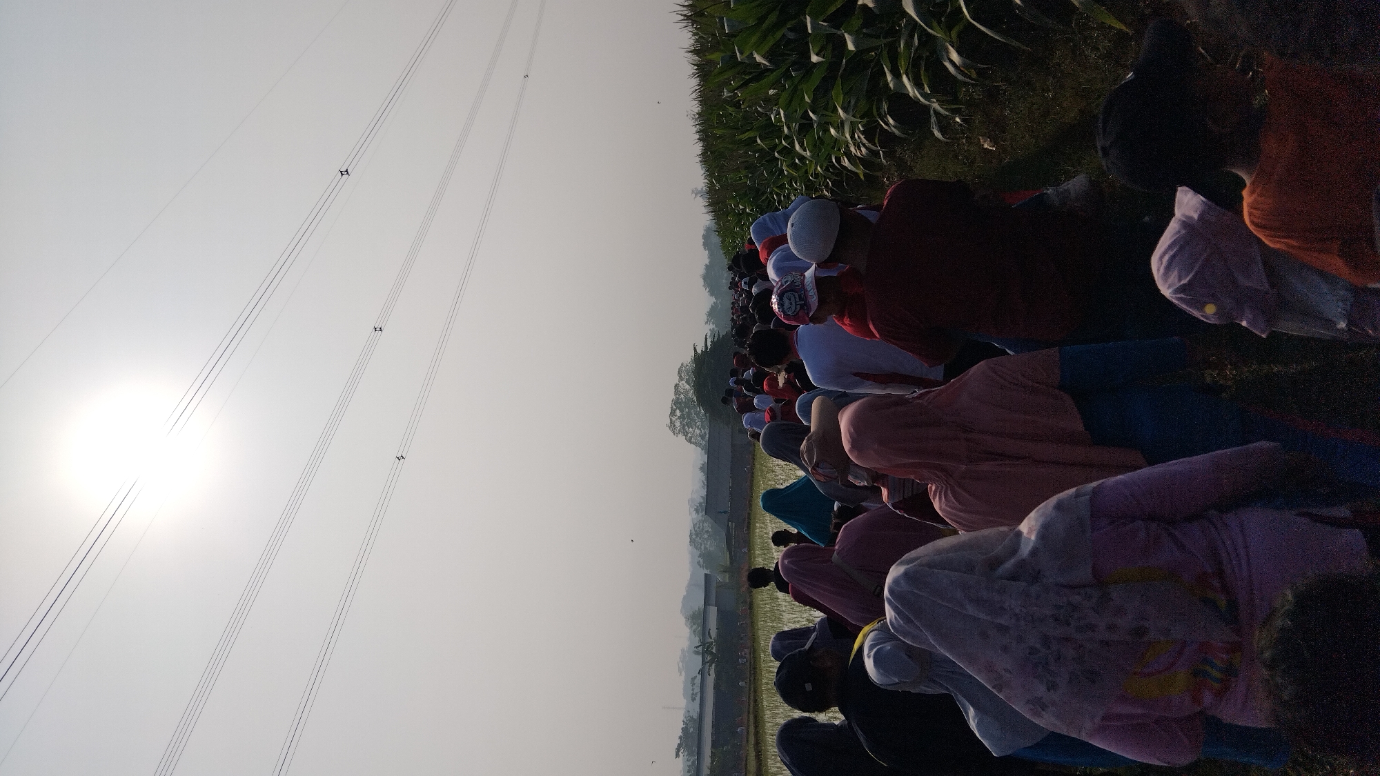 The image depicts a crowd of people walking along a path, bordered by a cornfield on the right and a rice paddy on the left. The sky is bright with the sun prominently featured, and power lines are visible overhead. The individuals are dressed in casual clothing, with many women wearing headscarves. The scene appears to be an outdoor event or procession, potentially in a rural area of Posong, Indonesia, given the agricultural landscape.