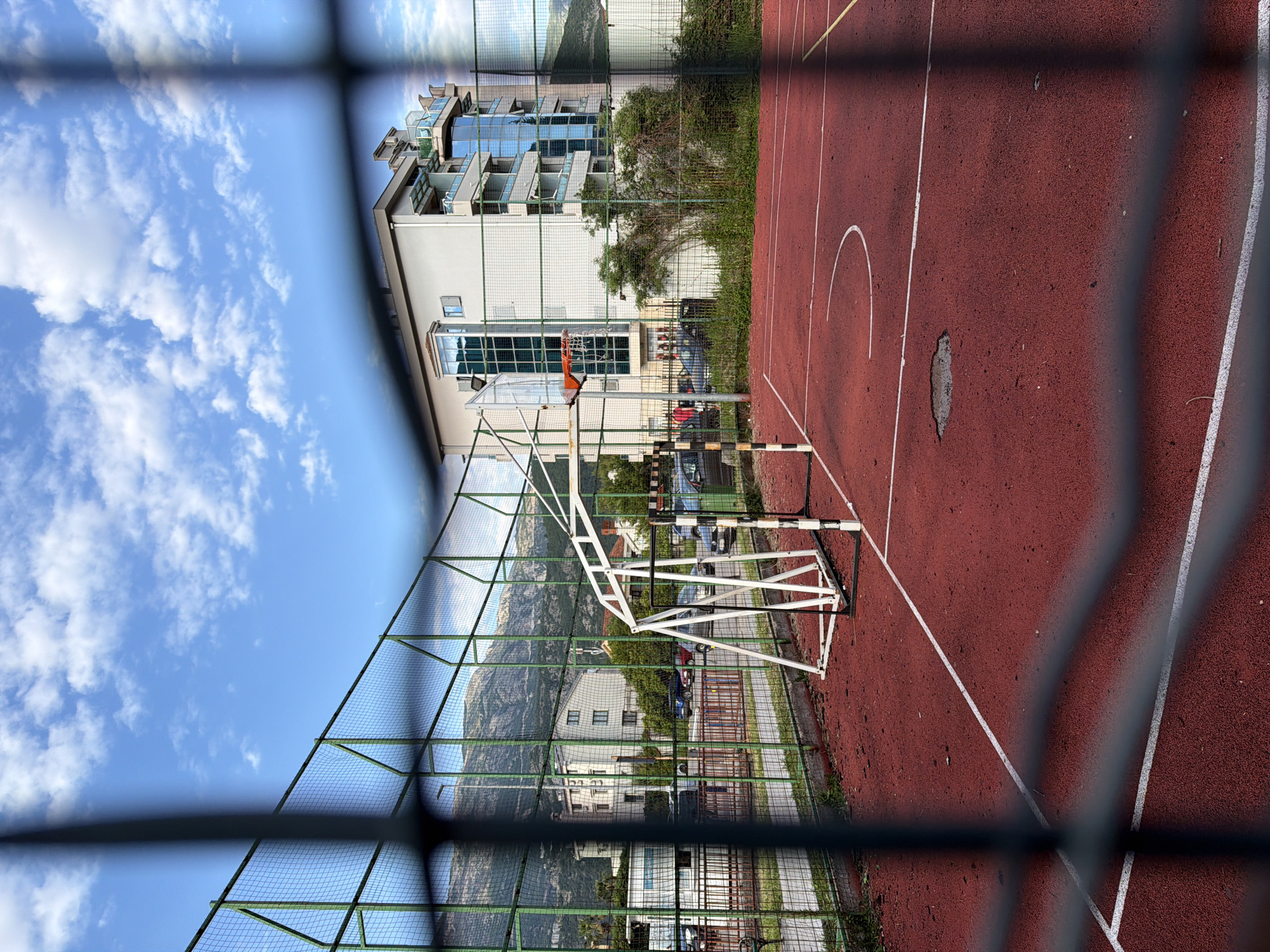 The image shows a basketball court in Budva, Montenegro, viewed through a chain-link fence. The court has a red rubber surface with white lines. A basketball hoop with an orange rim and white net is visible. A building with multiple floors is located in the background, and a mountain range is seen behind the building, partially obscured by the fence. The sky is blue with scattered white clouds. A sidewalk and greenery separate the basketball court from the building. There are cars parked along the street visible behind the fence.