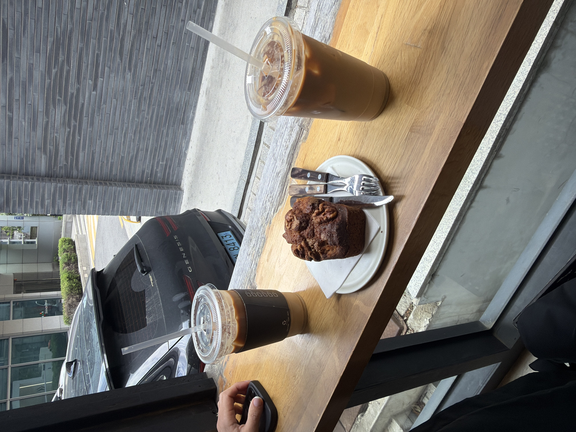 A wooden table near a window holds an iced coffee, a plate with a piece of cake, and cutlery. A second iced coffee and a hand holding a mobile phone sit closer to the window. Through the window, a black car with the word "GENESIS" visible is parked outside a building. The license plate reads "8413". The image appears to be taken in Seoul, South Korea, as the sign "Shimone Dental" is visible in the window.