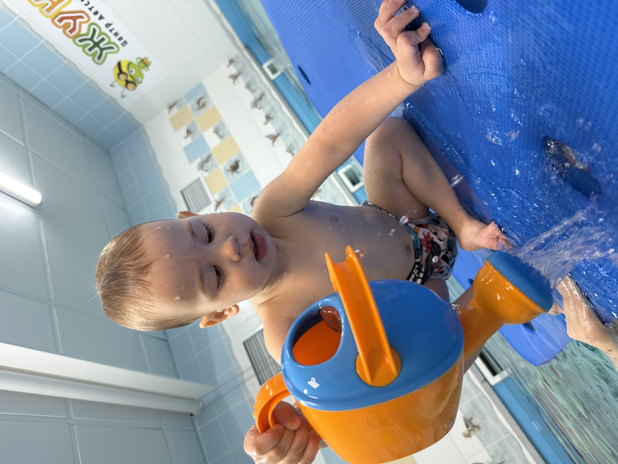 A toddler is playing with a blue and orange watering can in a pool at a swimming center. The swimming center is likely located in Rusanovo, Russia, given the sign in the background that reads "Центр Детск" (Children's Center) and the spelling of the center's name. The child is positioned on a blue floating mat in the water. The pool is surrounded by blue and white tiled walls. The child is fair-skinned and has short blond hair with water droplets on his head and body. The child wears a printed swim diaper. The watering can is being used to pour water into the pool.