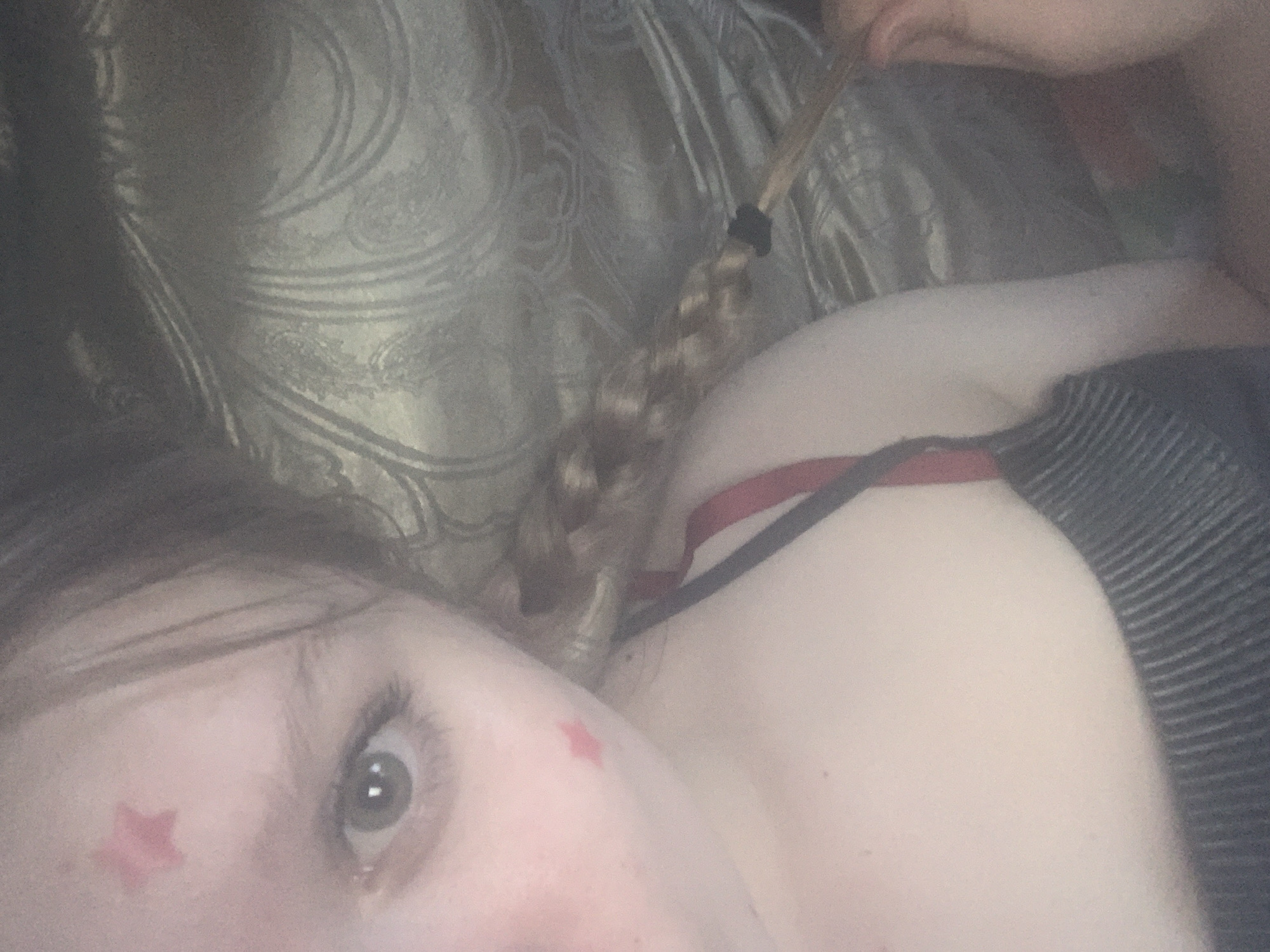 The image is a close-up, high-angle shot featuring a person lying down, possibly on a bed or sofa. The subject's left eye and part of their face, chest, and shoulder are visible in the lower and right sections of the frame. A small red star-shaped sticker or temporary tattoo is affixed to their left cheekbone, below the eye.
The person has light skin and features brown hair, some of which is styled in a braid secured with a black elastic band, visible towards the top-center. A hand is partially visible near the braided hair. The subject is wearing what appears to be a dark, possibly black, striped garment on their right shoulder, with a red bra strap visible beneath it.
The background consists of a textured fabric, likely a pillow or bedding, featuring a subtle, ornate pattern. The lighting is dim, suggesting an indoor setting with low ambient light. The person's expression is neutral, looking directly at the camera. There is no visible text in the image.