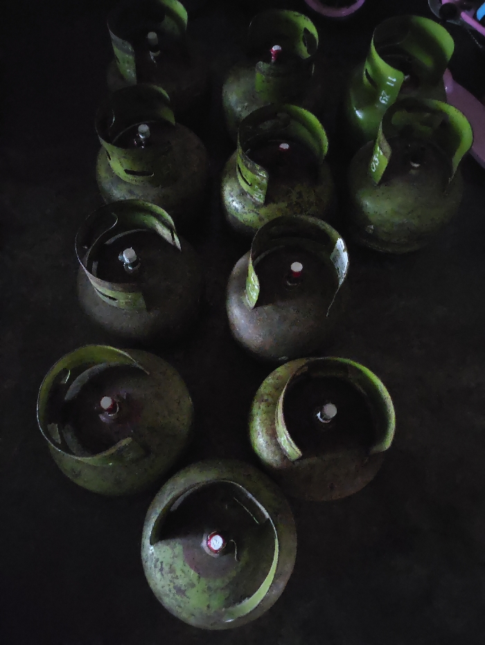 The image captures a collection of approximately ten green liquefied petroleum gas (LPG) cylinders, commonly used for cooking gas, arranged on a dark, textured surface. The cylinders are depicted from a top-down perspective, showcasing their circular protective guards and central valves. Many of the cylinders display visible signs of wear, such as scratches, scuffs, and dirt, indicating regular use.
No people are present, and there is no specific activity taking place; the cylinders appear to be stationary, possibly stored or awaiting distribution. The setting suggests an indoor space or a shaded outdoor area, characterized by dim, ambient lighting that creates soft shadows and a somewhat subdued atmosphere. This scene is consistent with a storage or supply point for household gas in Selagai Lingga, Indonesia, where these green cylinders are a common sight.
Notable details include text visible on some of the cylinders' protective guards. On a cylinder in the upper right, the numbers "11 23" are faintly discernible. Another cylinder positioned towards the mid-right shows partial text, possibly "03/1" or "03/". These markings likely serve as identifiers such as manufacturing dates or batch numbers. The time of day cannot be determined due to the overall dim illumination. No emotions are conveyed by the inanimate objects.