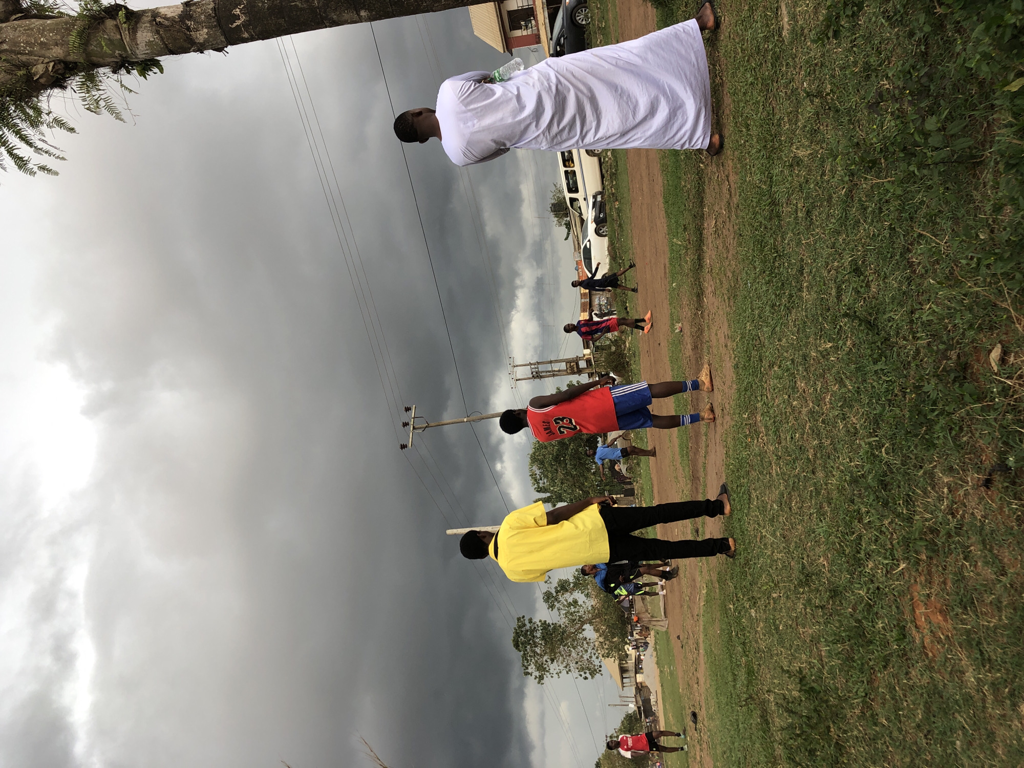 The image captures an outdoor scene under a heavily overcast, dark grey sky, suggesting impending rain or a storm. A large palm tree trunk with ferns is visible in the upper left corner, while utility poles and power lines crisscross the middle ground.
In the foreground, several individuals are present on a grassy and dirt area that appears to be an informal playing field or open space. One man, seen from behind, wears a long white robe (possibly a thawb or djellaba) and holds a plastic water bottle. To his left, another man, also viewed from behind, is dressed in a bright yellow shirt and black trousers.
Further to the left, a young man wears a red sports jersey with "JAMES 23" printed on the back, paired with blue shorts and blue and white striped socks. Several other young men or boys, some in blue and red sportswear, are scattered across the scene, either standing or in various stages of movement, suggesting they might be participating in or observing a sports activity, possibly football.
In the background, dirt roads or paths are visible, alongside simple buildings and parked vehicles, including a white van, indicating an urban or semi-urban setting. The ground consists of patches of green grass interspersed with bare earth.