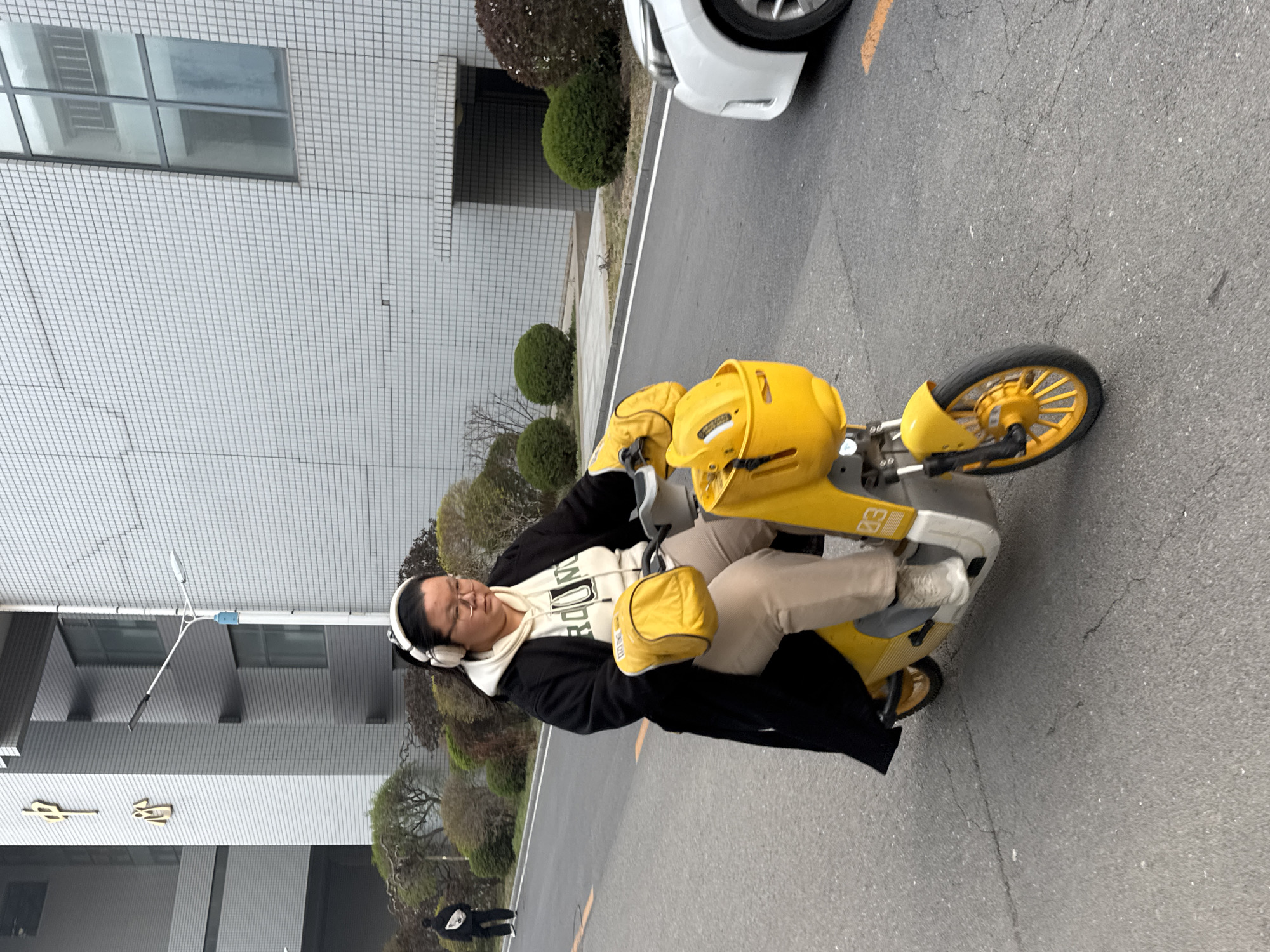 A woman is riding a yellow scooter on a paved road. She is wearing beige pants, white sneakers, a white hoodie, and a black jacket. She has headphones on. A white car is partially visible in the upper right corner. The background includes a building with a light gray tiled facade, trees, and bushes. A person in black clothing is walking in the background. A building with golden symbols is visible in the left. Location: possibly Zhongtun, China.