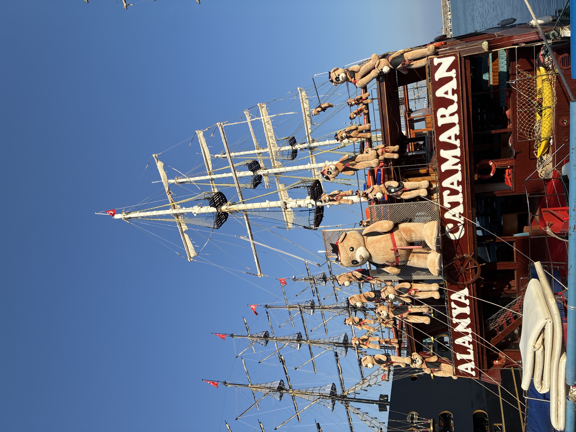 The image shows the stern of a large, wooden, multi-masted ship named "ALANYA CATAMARAN" docked in the harbor of Alanya, Turkey. The ship's name is prominently displayed in white lettering against a dark red background. Numerous large, stuffed teddy bears, some wearing hats and vests, are positioned along the railings and decks of the ship, with some hanging from ropes. The ship has multiple decks and rigging characteristic of a sailing vessel. In the background, another large dark-colored ship is partially visible, and the clear blue sky dominates the upper portion of the image. The sea is visible to the right. There are Turkish flags flying on the masts of the ship. The overall scene suggests a tourist attraction or charter vessel.