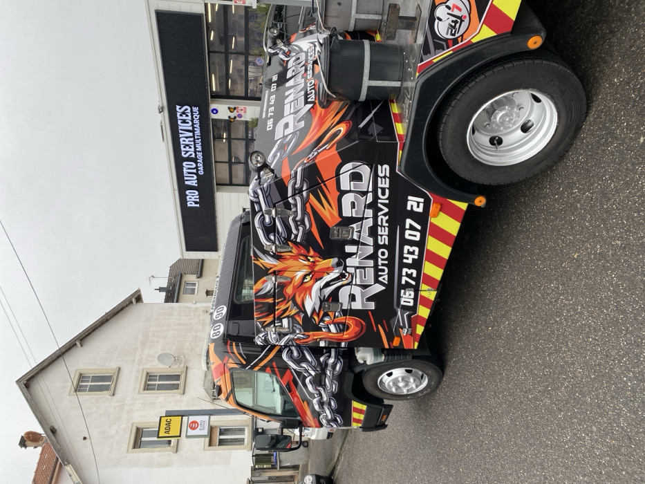 A customized tow truck, branded "RENARD AUTO SERVICES," is parked on an asphalt road in Bischheim, France, under an overcast sky. The truck features a prominent graphic of an orange fox head amidst black and orange flame-like patterns and chains on its side. White text displays "RENARD AUTO SERVICES" and a phone number "06 73 43 07 21." Red and yellow diagonal stripe reflective markings are visible along the lower body, and a "24/7" logo is present near the rear wheel well. The vehicle has dual rear wheels, searchlights mounted on the top rear of the cab, and two circular speed limit stickers (80 and 90) on the rear of the cab roof. A dark bucket is placed on the flatbed section behind the cab. To the left, a light-colored multi-story building, possibly residential, with multiple windows and a satellite dish, stands alongside a pole bearing a yellow ADAC sign and an "E PRO REPAR" sign. To the right, a "PRO AUTO SERVICES GARAGE MULTIMARQUE" facility is visible with large glass garage doors and a dark grey sign board. No individuals are present.