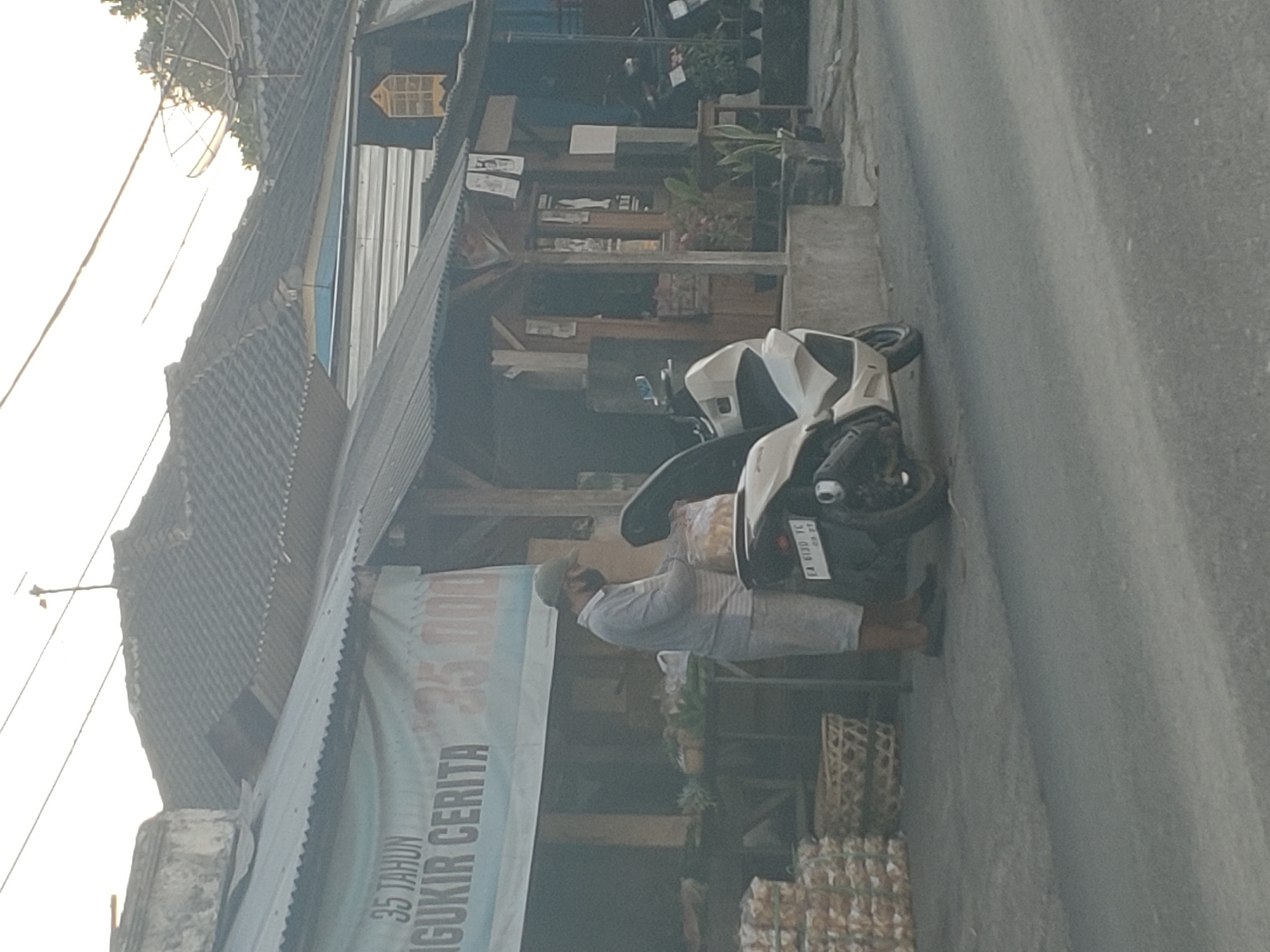 A man wearing a grey long-sleeved shirt, white shorts, a green cap, and a black face mask stands on a paved street in Kembang Kerang, Indonesia. He is packing goods into a clear plastic bag. To his right, a white scooter is parked. In front of the man, stacked baskets and produce are visible on a low table. Behind him, a building with a corrugated metal roof is visible, featuring a banner that reads "35 TAHUN NGGUKIR CERITA" and "35.000". Other buildings and vegetation are also present in the background. The lighting suggests it is either early morning or late afternoon.