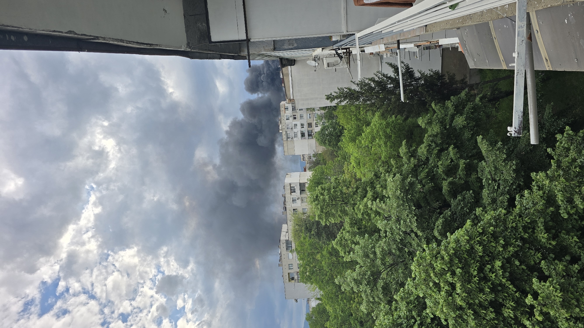 The image, taken in Sofia, Bulgaria, shows thick, dark smoke billowing into a cloudy sky. Adjacent to the smoke are multi-story apartment buildings, lined with trees in full leaf. The photograph appears to be taken from an elevated position, likely a balcony.