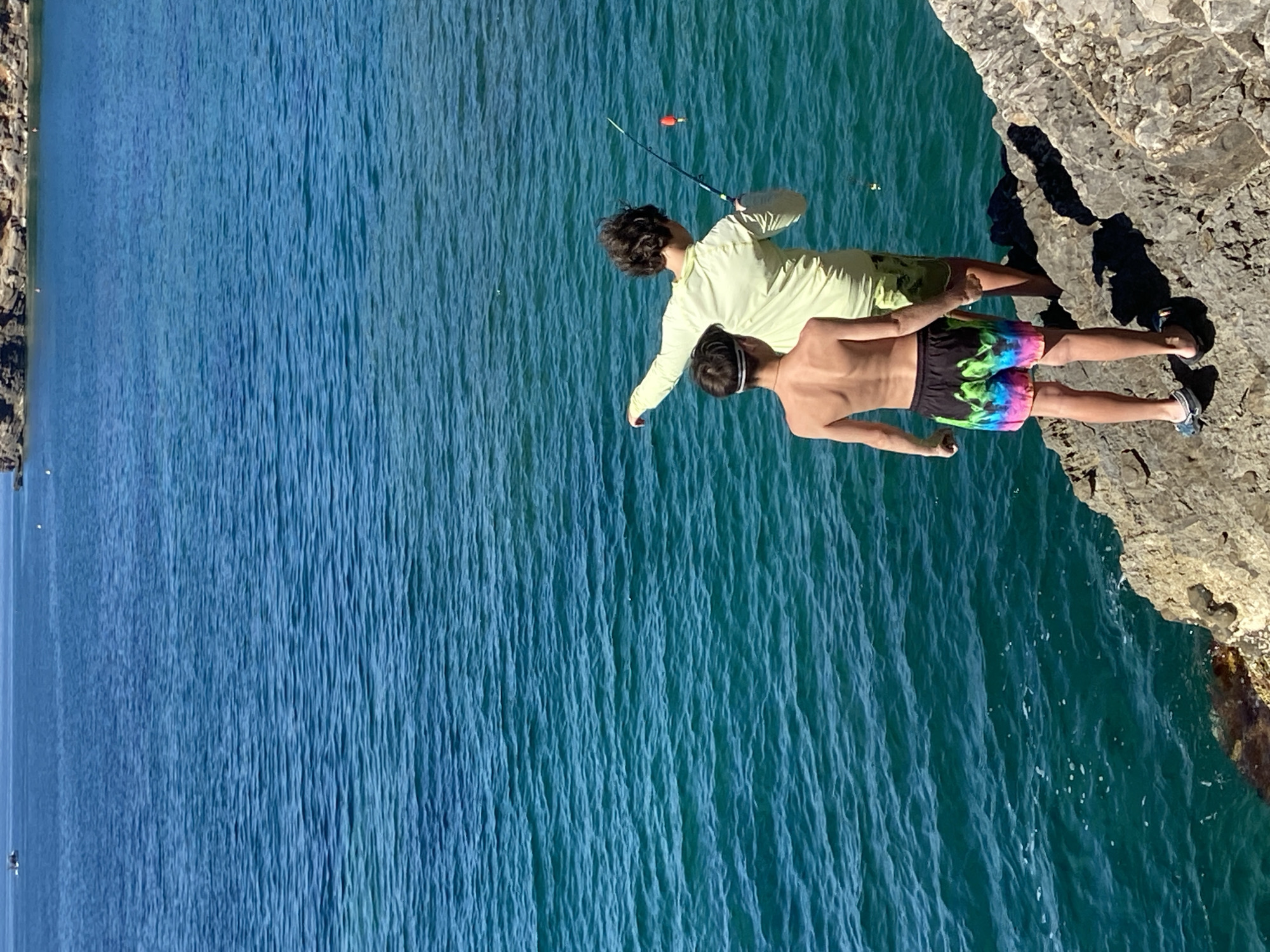 Two young males are standing on a rocky shoreline and fishing in the clear, blue-green waters of the Adriatic Sea, likely near Budva, Montenegro. The male in the foreground is facing away from the camera, wearing multi-colored swim shorts. The male behind him, also facing away, is wearing a light yellow long-sleeved shirt and green patterned shorts, holding a fishing rod with a red and orange float. The water is calm with small ripples, and in the distance, a rocky coastline is visible, with faint hints of other boats or structures on the horizon. The scene is illuminated by bright sunlight.