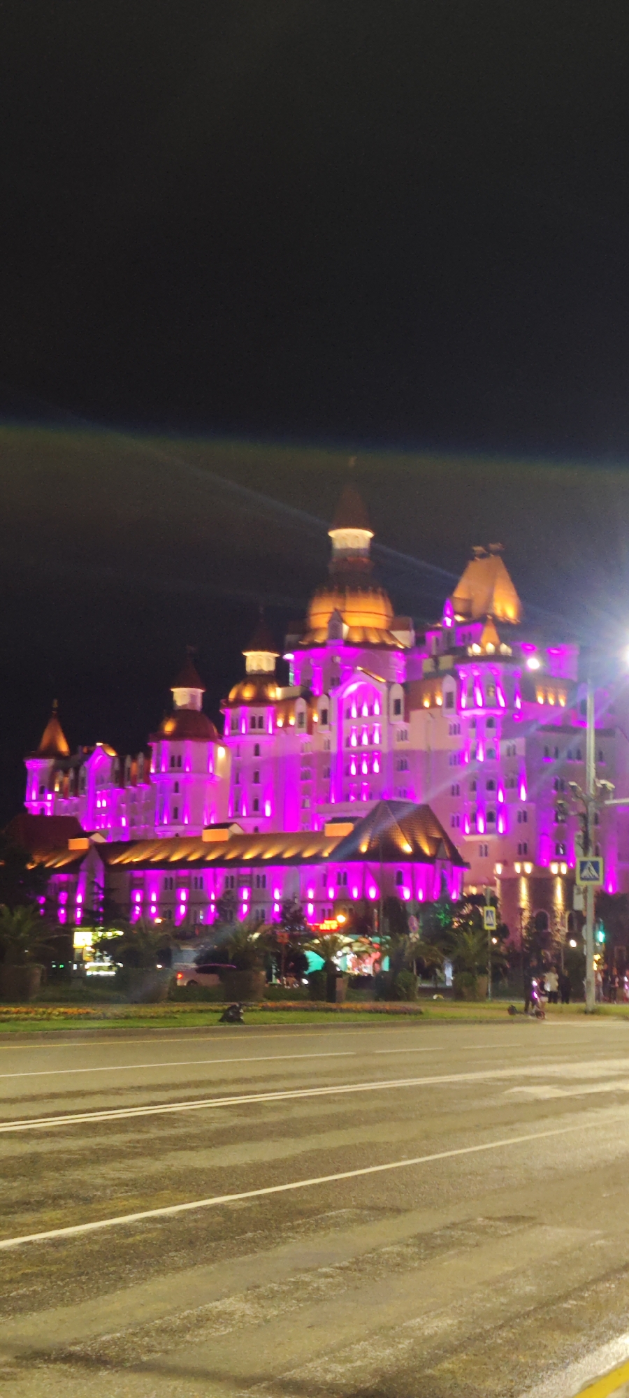 At night, a large building with multiple towers is illuminated with pink and orange lights. The structure, likely a hotel or resort, is located in the city of Sirius, Russia, as indicated by street signs. A wide, wet asphalt road runs in front of the building, with faint white lines marking lanes. Vegetation, including trees and flower beds, is visible in the median between the road and the building. In the background, beyond the building, the sky is black. A streetlight on the right side of the image adds additional illumination, and a faint lens flare is visible at the top left. The focus is on the building and its colorful lighting. A couple of people are visible near a crosswalk.