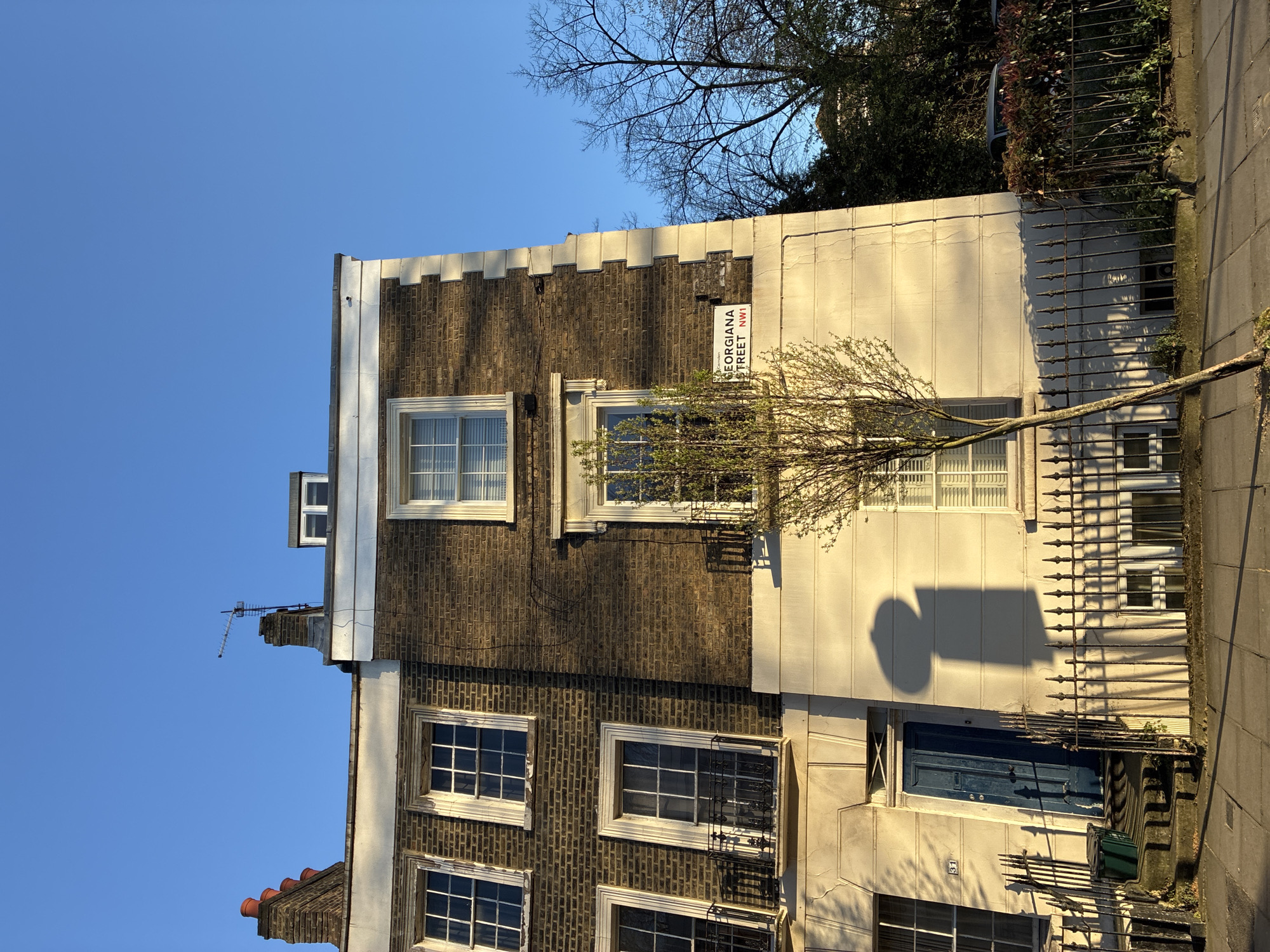 The image shows a building in London, United Kingdom. A street sign reads "Georgiana Street, NW1". The building has a dark brick exterior on the left, and a light beige exterior on the right. There are multiple windows with white frames, and a blue door on the right side of the building. A small tree is partially obscuring one of the windows. An antenna is visible on the roof of the building. A wrought-iron fence borders the building. The sky is blue.