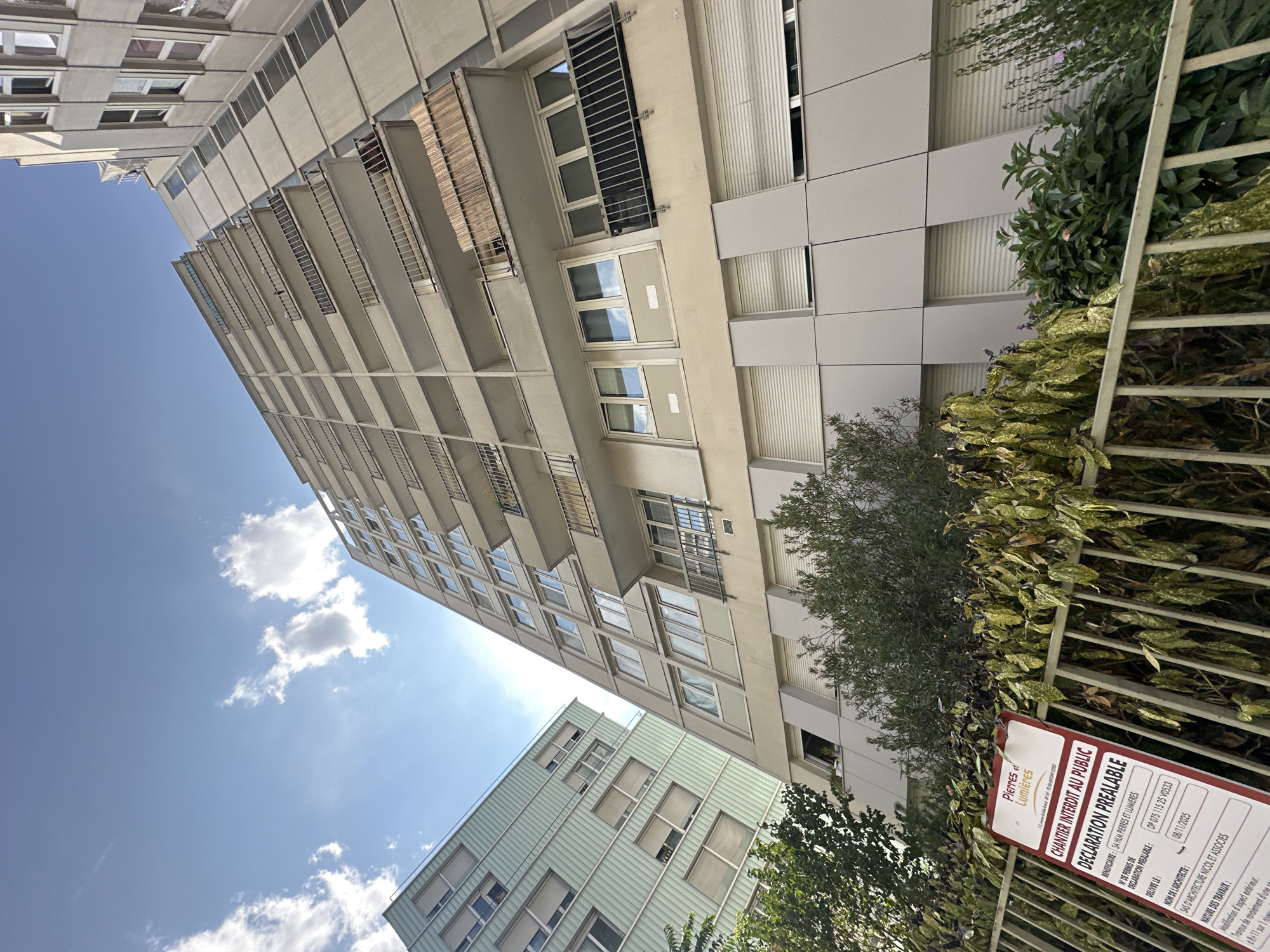 The image shows a low-angle view of two apartment buildings, one taller and beige, the other shorter and light green. The beige building has numerous balconies with metal railings and wooden slats on some. The windows on both buildings have roller shutters. In the foreground, a metal fence with a sign is visible. The sign is red and white and reads "CHANTIER INTERDIT AU PUBLIC DECLARATION PREALABLE" which translates to "CONSTRUCTION SITE FORBIDDEN TO THE PUBLIC PRIOR DECLARATION." Below this, there is text in French indicating project details, including a project permit number and a deadline of 09/11/2025. The sky above is blue with scattered white clouds. Trees and shrubs are present in front of the fence. The architectural style suggests a mid-to-late 20th-century urban development, typical of residential areas in Paris, France. The scene appears to be a typical urban construction site.