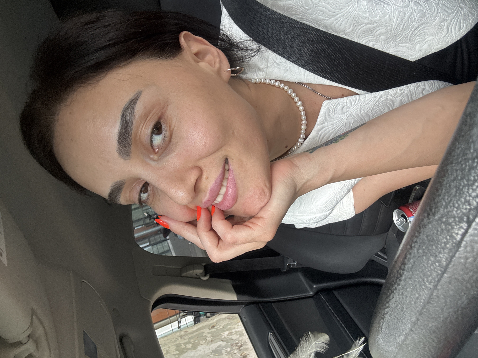 A medium shot captures a woman in the passenger seat of a car, likely in Tbilisi, Georgia, given the background architecture and common car interiors found there. She is leaning her head on her hand, with her left hand fingers gently touching her cheek and mouth. Her eyes are looking towards the camera with a slight smile. She has dark hair and is wearing makeup, including bright orange nail polish. A pearl necklace adorns her neck, and a silver necklace is visible underneath. Her outfit is a white textured top with a black seatbelt across her chest. Inside the car, a can of Coca-Cola is visible on the center console. Outside the car window, a tree and buildings can be seen, suggesting an urban setting. The overall impression is a casual, self-captured moment.