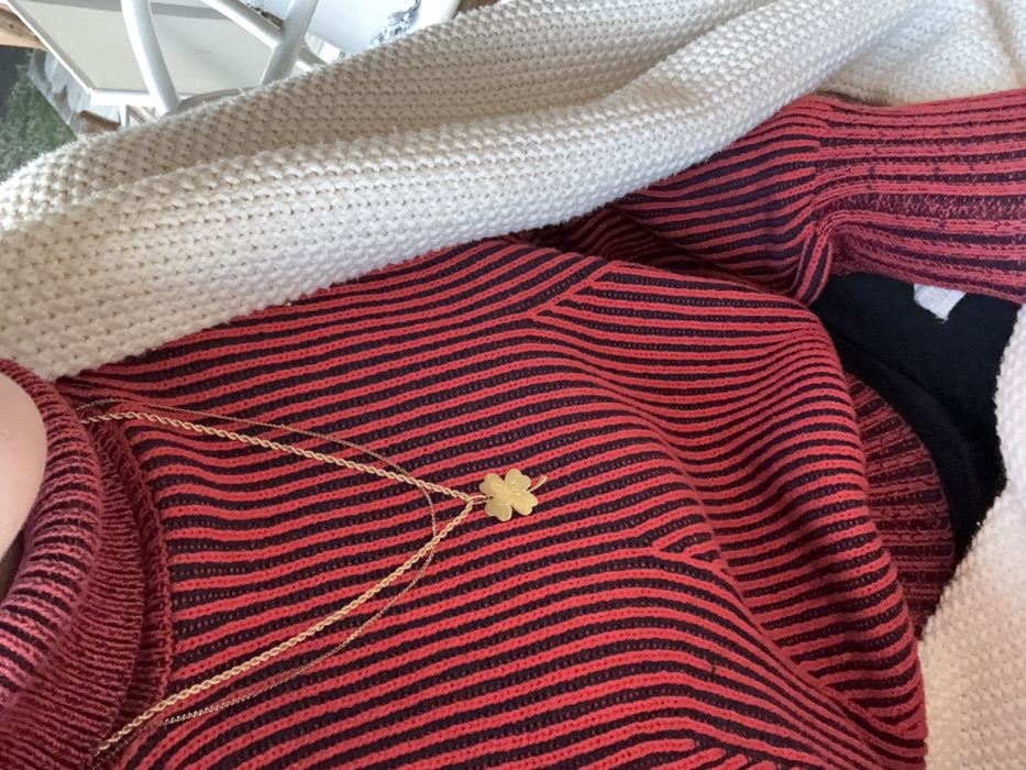 The image shows a red and black striped sweater with a gold clover necklace on top. The sweater is partially covered by a white knit blanket or garment. Part of a white chair is in the upper left corner. Hints of black and white fabric are visible underneath and to the right of the red sweater.
