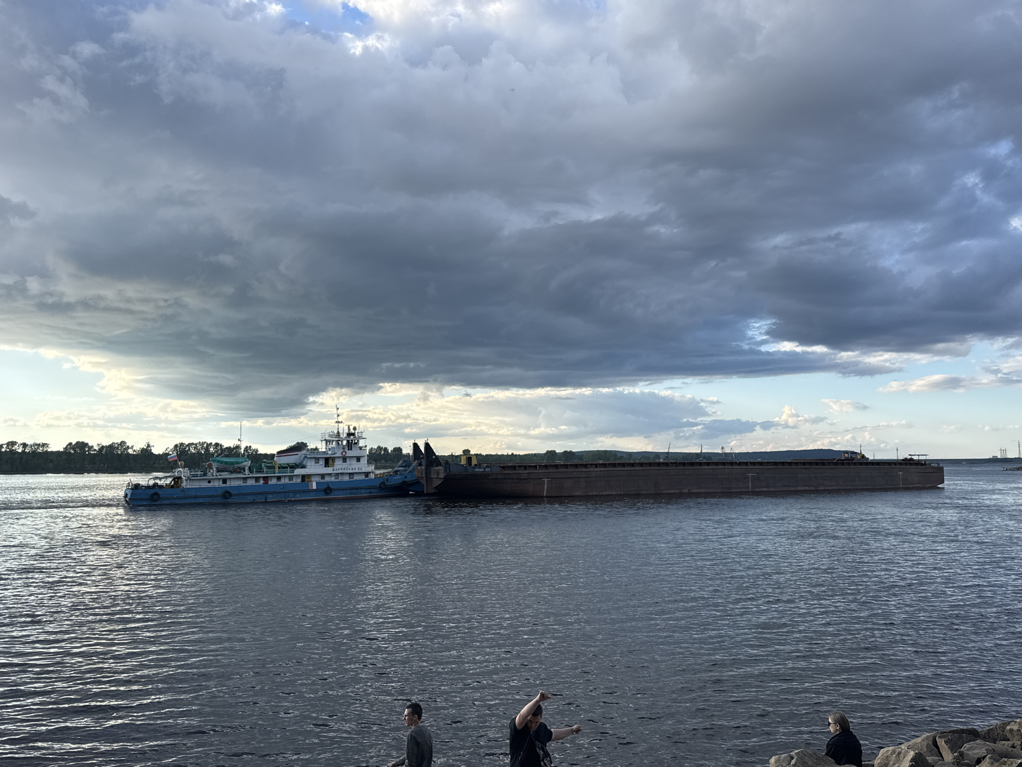 The image captures a wide river with a tugboat towing a barge under a dramatic, cloud-filled sky. In the foreground, three individuals are visible on a rocky shore, two of whom are facing away from the camera. The tugboat, painted blue and white, has Russian lettering on its side, "ДМПДЯНЬ 54". The barge is long and flat, with a dark, weathered surface. The opposite bank of the river is lined with trees, suggesting a natural landscape. In the far distance, industrial structures, possibly related to the city of Chaikovsky, are faintly visible, including what appears to be a power line tower. The overall impression is one of a working river scene with a hint of approaching weather.