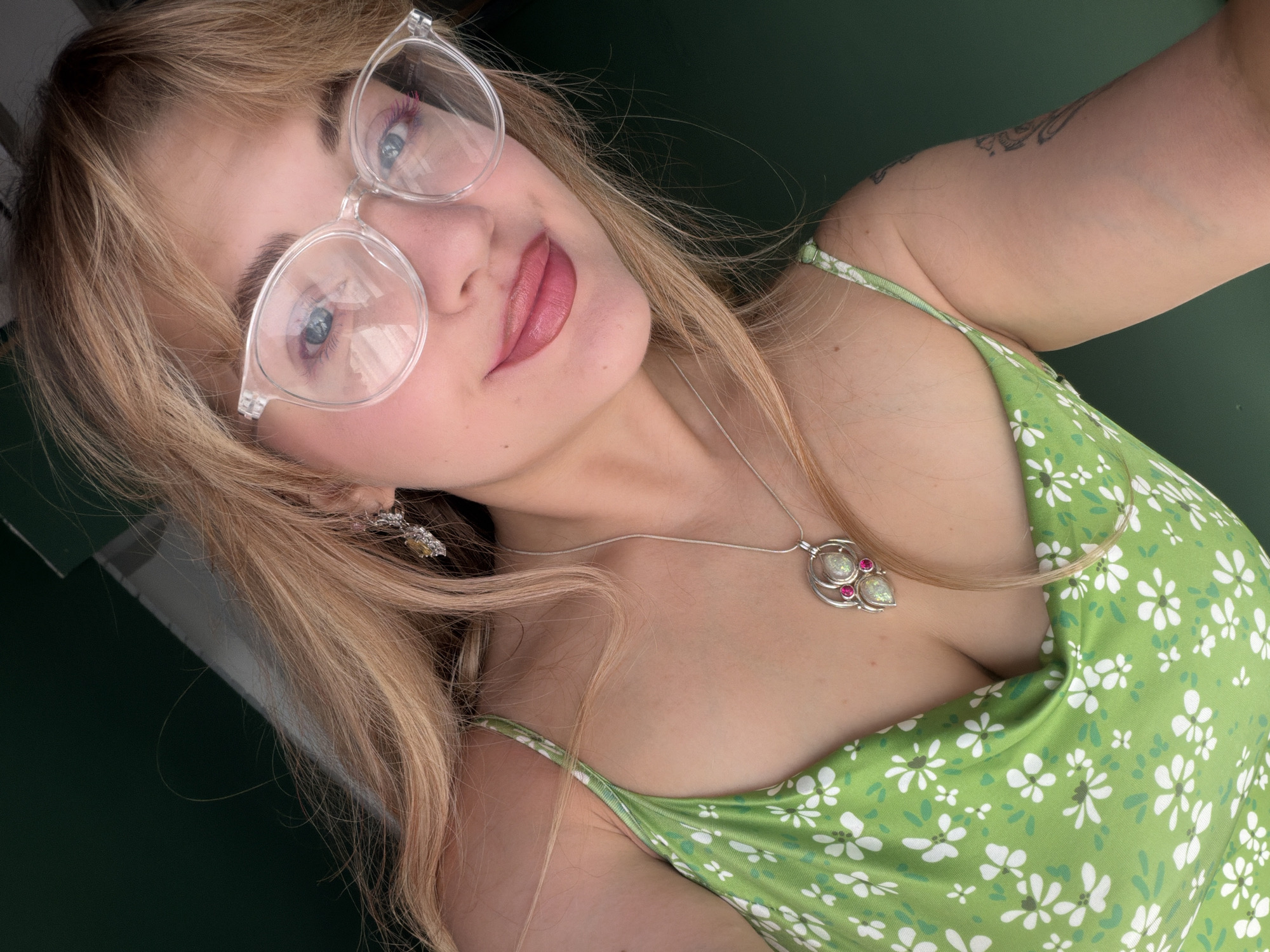 The image is a selfie of a woman with blonde hair wearing clear-framed glasses, a green floral dress, and a silver necklace. She has a tattoo on her left arm. Her tongue is slightly out, and she is looking up at the camera. The background is a dark green. There is no information about the location being Большая Шильна, Russia.