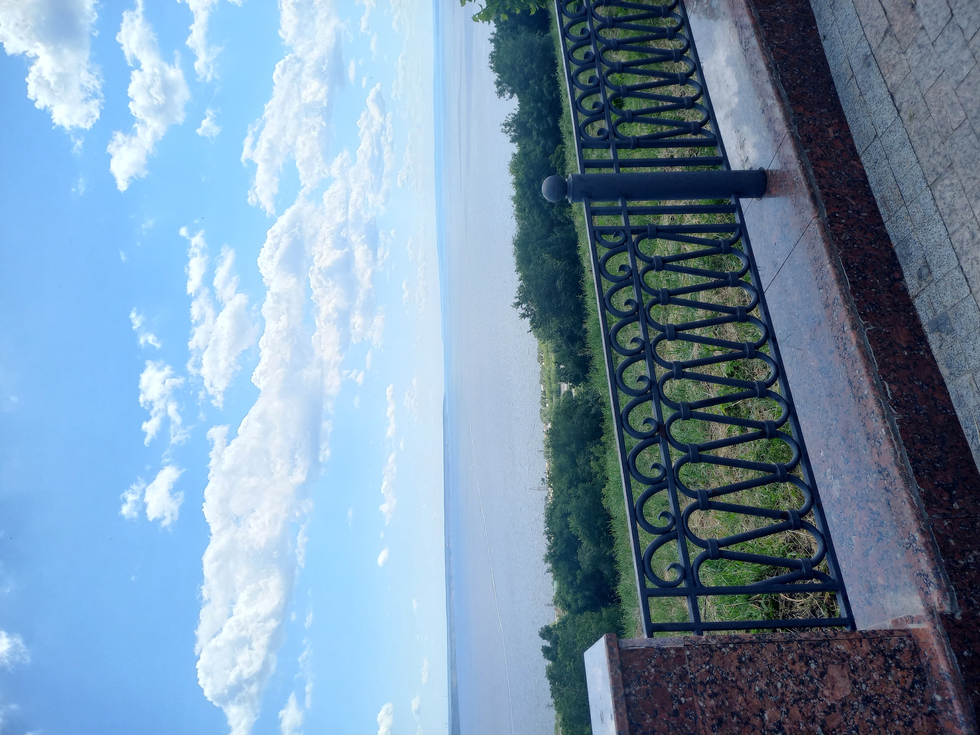The image shows a scenic overlook of the Volga River in Ulyanovsk, Russia, on a sunny day with a partly cloudy sky. In the foreground, there is a decorative wrought iron fence with a granite base and pillar, suggesting a public viewing area. Beyond the fence, a dense line of green trees separates the overlook from the expansive blue water of the river. The river stretches to the horizon, where it meets a hazy sky. The overall impression is one of tranquility and natural beauty.