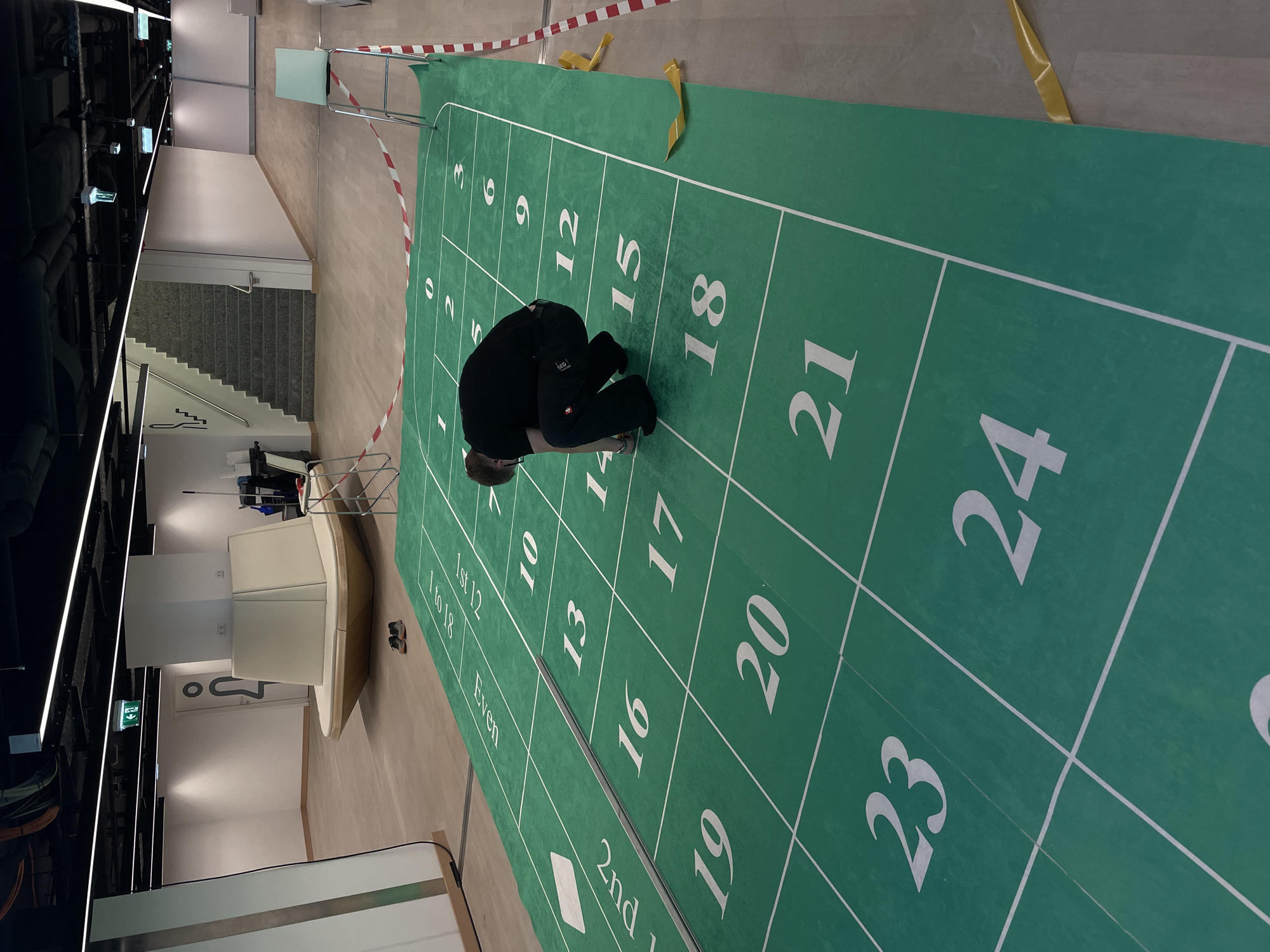 The image depicts an indoor space featuring a large, green hopscotch court with numbered squares ranging from 0 to 24, as well as additional spaces labeled "1st 12," "1 to 18," "Even," and "2nd 12." A person wearing black clothing is crouched on the court, near the squares marked "14" and "17." The court is bounded by a red and white barrier tape in some sections, and yellow tape in others.
In the background, the scene includes a modern interior with stairs, furniture, and architectural elements. The presence of the hopscotch court suggests a recreational or play area. The location seems to be a professional setting. The specific city or location within the city is not discernible from the provided image.