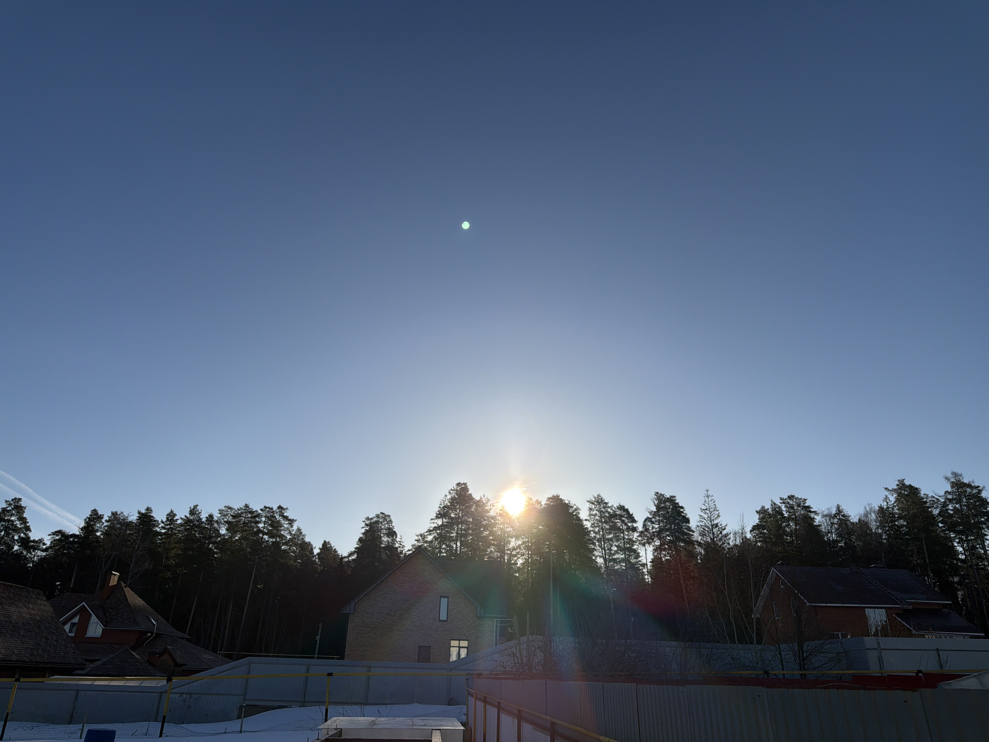 The image shows a daytime scene in what appears to be a residential area. The sky is a clear, light blue, with a bright spot suggestive of a lens flare or the sun itself near the center. Below the sky, a line of trees obscures the horizon. The sun is positioned behind the trees, creating a visible lens flare.
In the foreground, several houses can be seen partially obscured by the trees. To the left and right are glimpses of rooftops and walls of houses that seem to be made of brick or siding. A metal fence runs across the lower portion of the frame. There is snow visible in the lower-left quadrant, suggesting a winter or early spring setting.
The image offers no clear evidence to confirm that this is specifically Chaikovsky, Russia. Therefore, based only on the image, the description cannot be more specific. There are no people or distinct objects easily discernible. The action is simply the state of being in this environment, a residential area in a place with trees and snow.
