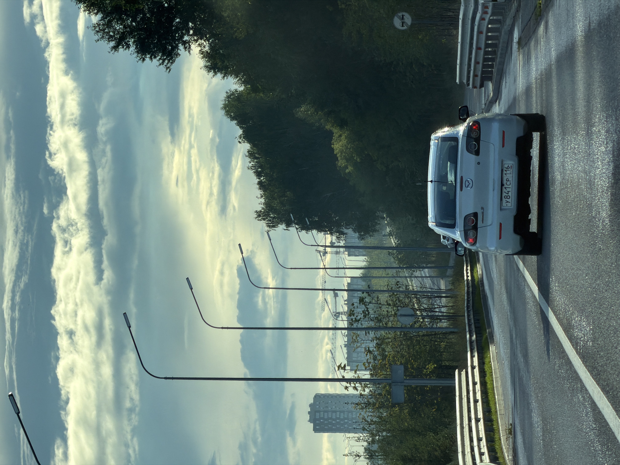 A white Mazda 3 car is driving on a wet asphalt road. The car has a Russian license plate that reads "Y841CP 116". The road is lined with streetlights and guardrails. In the background, there are trees, buildings, and cranes, suggesting an urban environment. The sky is cloudy. Based on the license plate, the location is Kazan, Russia.
