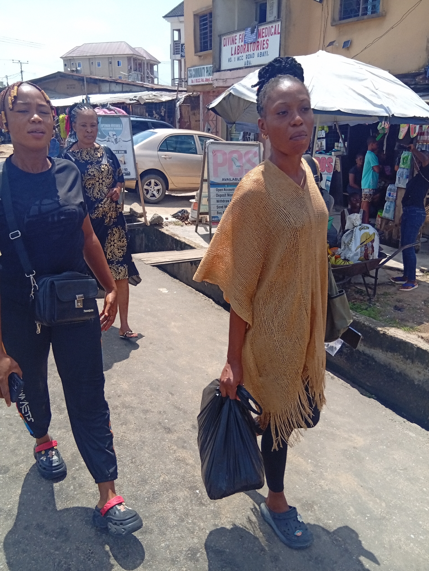 The image captures a street scene in Aba, Nigeria. Three women are the primary subjects: one woman wears a black t-shirt, track pants, and decorated clog-style shoes, carrying a small purse. Another woman in a floral-patterned dress and sandals is behind her. The third woman, in the foreground, wears a tan knitted poncho, leggings, and blue clog-style shoes, holding a black plastic bag.
In the background, there is a beige-colored sedan, a building with signage for "DIVINE FAV MEDICAL LABORATORIES NO. 1 MCC ROAD ABAYI. ABA," and a POS sign indicating available services. A vendor operates under a white umbrella with a wheelbarrow nearby. The environment seems to be a bustling street with pedestrians and small businesses. The weather appears sunny.