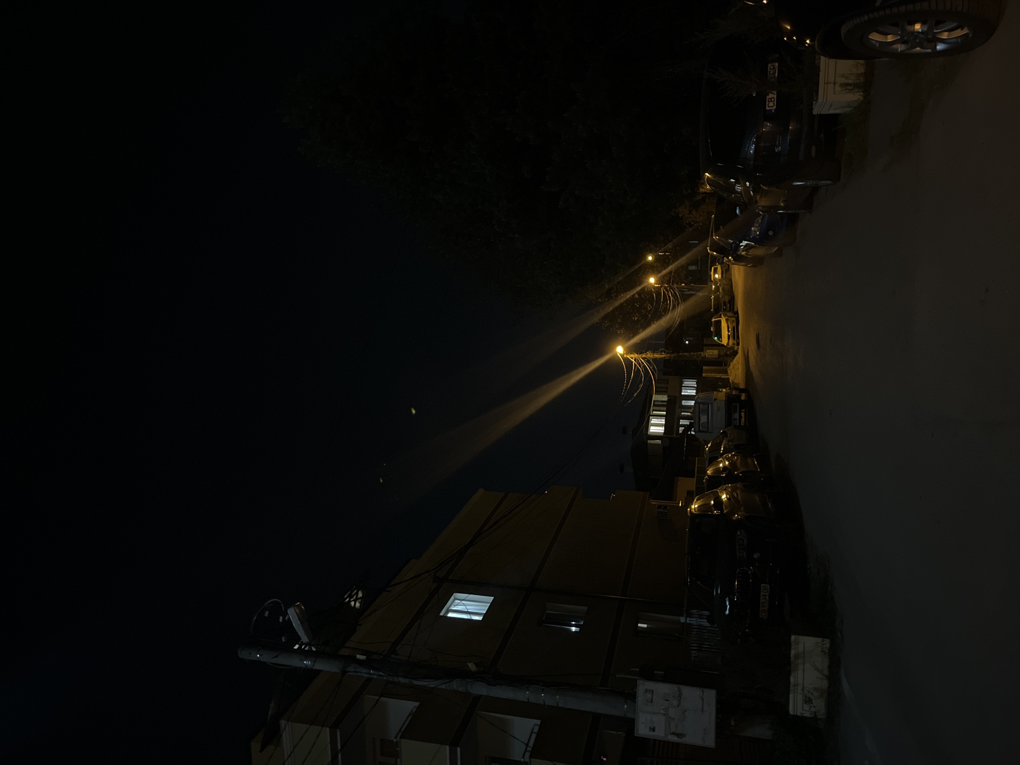 The image captures a nighttime scene on an urban street, likely in Sofia, Bulgaria, as indicated by the context and visible license plate prefixes. The perspective is oriented 90 degrees counter-clockwise, showing a street with parked cars on the right and a building on the left.
Several vehicles are parked in a row along the right side of the paved road. Their dark bodies are partly illuminated by the bright, warm light from streetlights lining the street. These streetlights are a prominent feature, casting intense, starburst-like lens flares that radiate across the scene.
On the left, a multi-story building stands, with at least two of its windows glowing brightly from internal lights, suggesting occupancy. In the lower left foreground, a utility pole is visible, laden with wires and some equipment, extending upwards towards the building. The upper left and background areas are dark, revealing the night sky and the silhouettes of trees or dense foliage.
The scene depicts a quiet and still urban environment after dark, with no people or specific activities taking place. The time of day is clearly night, characterized by the deep darkness and reliance on artificial lighting.
Visible text includes parts of car license plates. One plate clearly shows "CB 74..." and another "CB 80...", consistent with registration codes for Sofia, Bulgaria.
